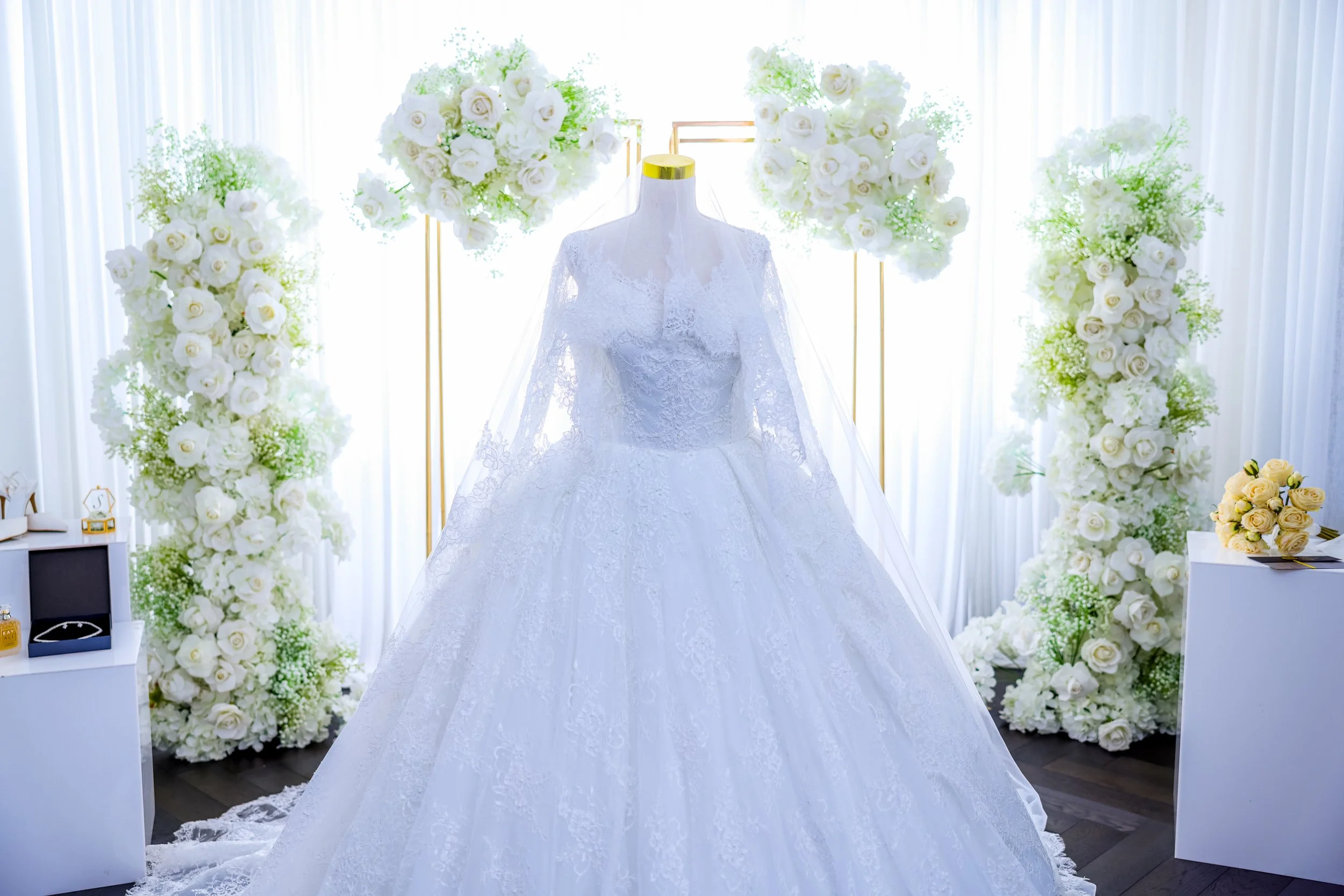 A white wedding dress displayed on a mannequin, surrounded by white and yellow flowers in an elegant setting.