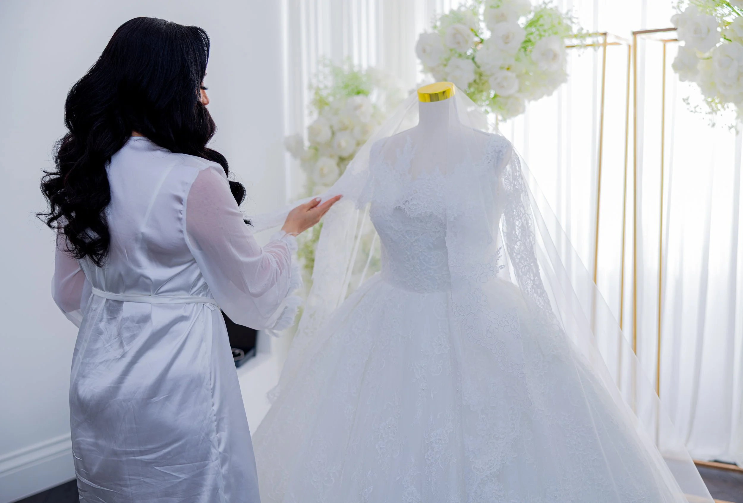 A woman with long black curled hair wearing a white satin robe is examining a wedding dress on a mannequin, in a bright room with white curtains and floral decorations.