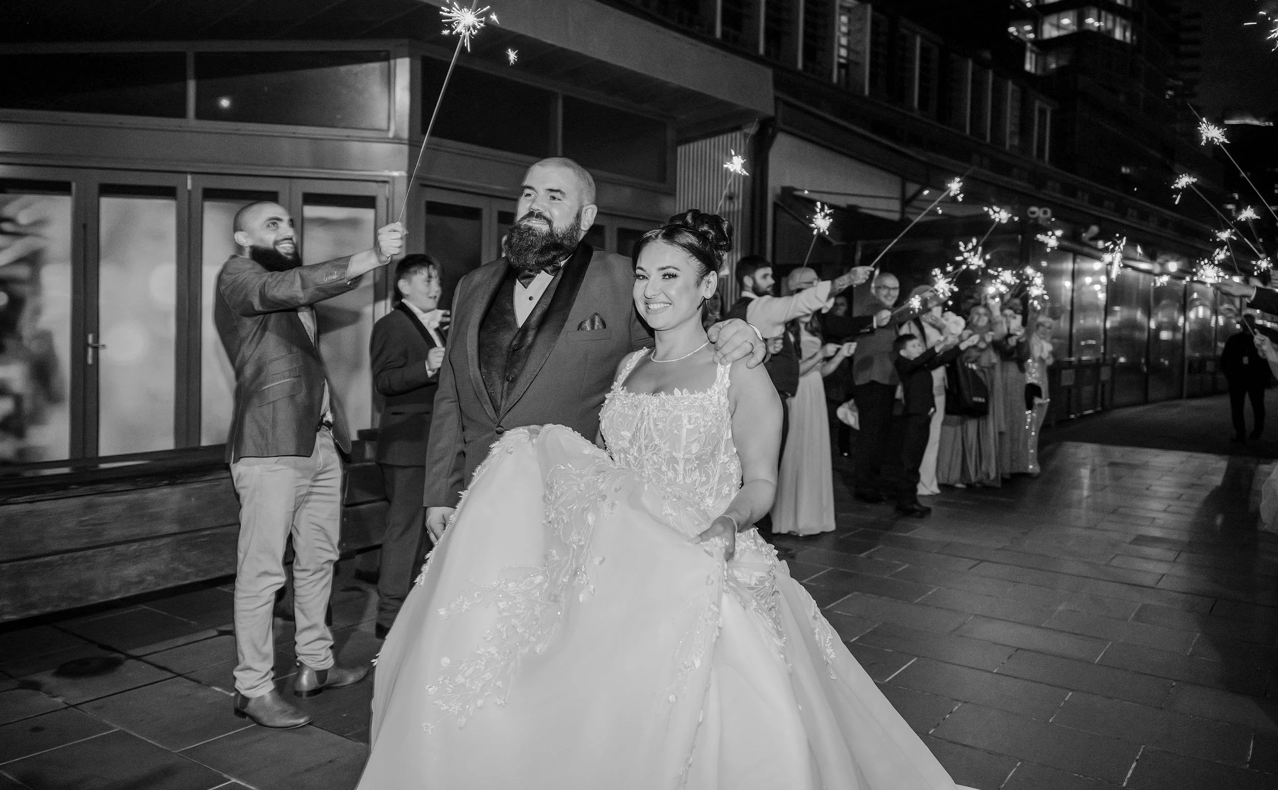 Wedding couple smiling, surrounded by guests holding sparklers at night.