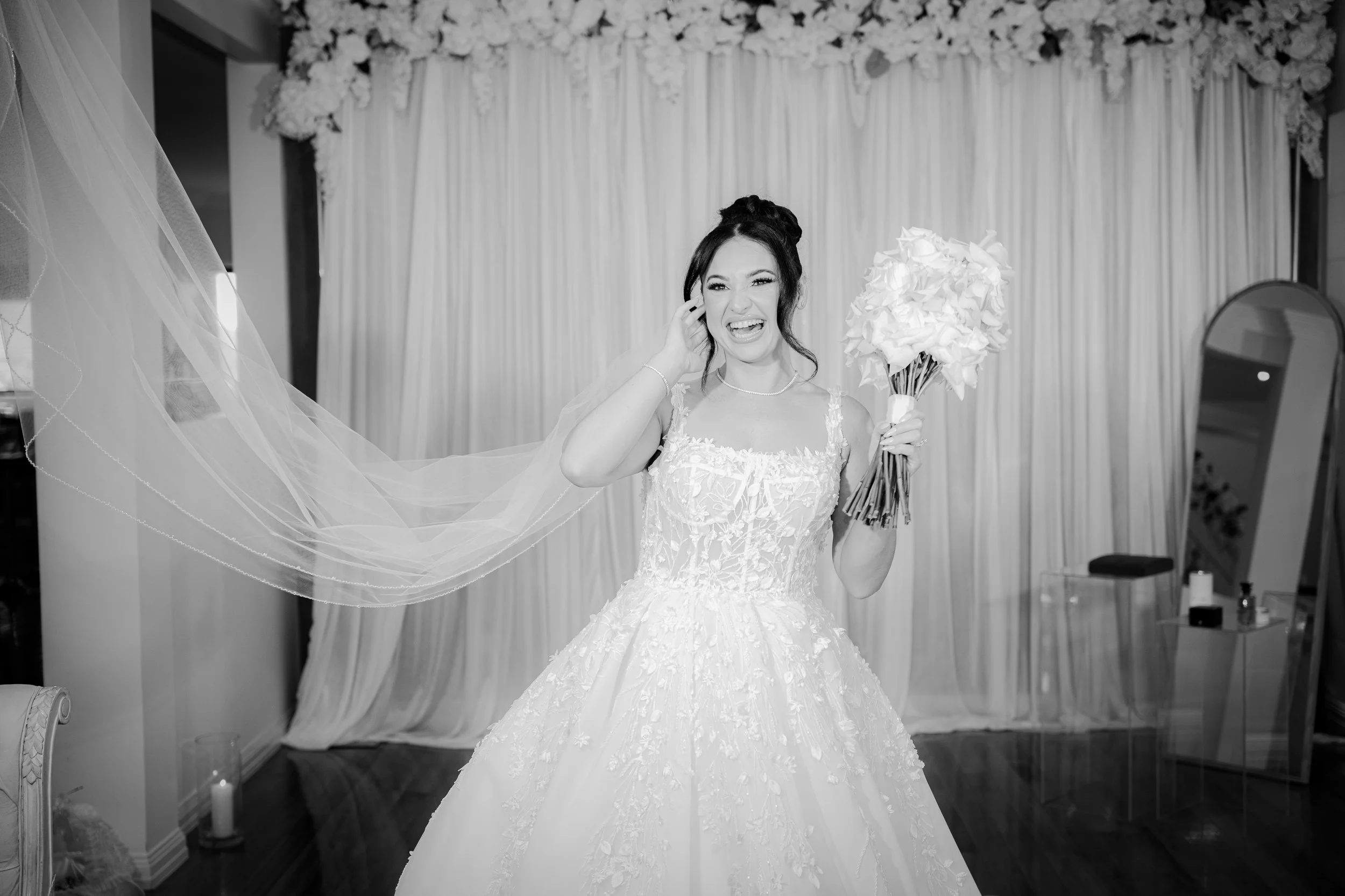 A bride in a wedding dress holding a bouquet of flowers, smiling and winking, in a decorated room with curtains and floral arrangements.