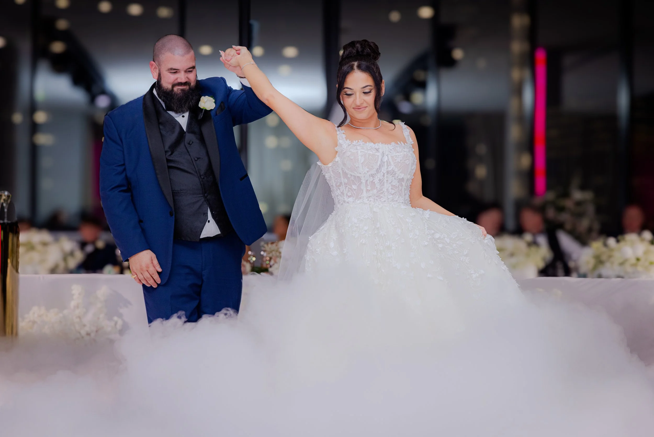 A bride and groom dance at their wedding reception, with the groom wearing a blue tuxedo and the bride in a white wedding gown. There is fog on the floor.