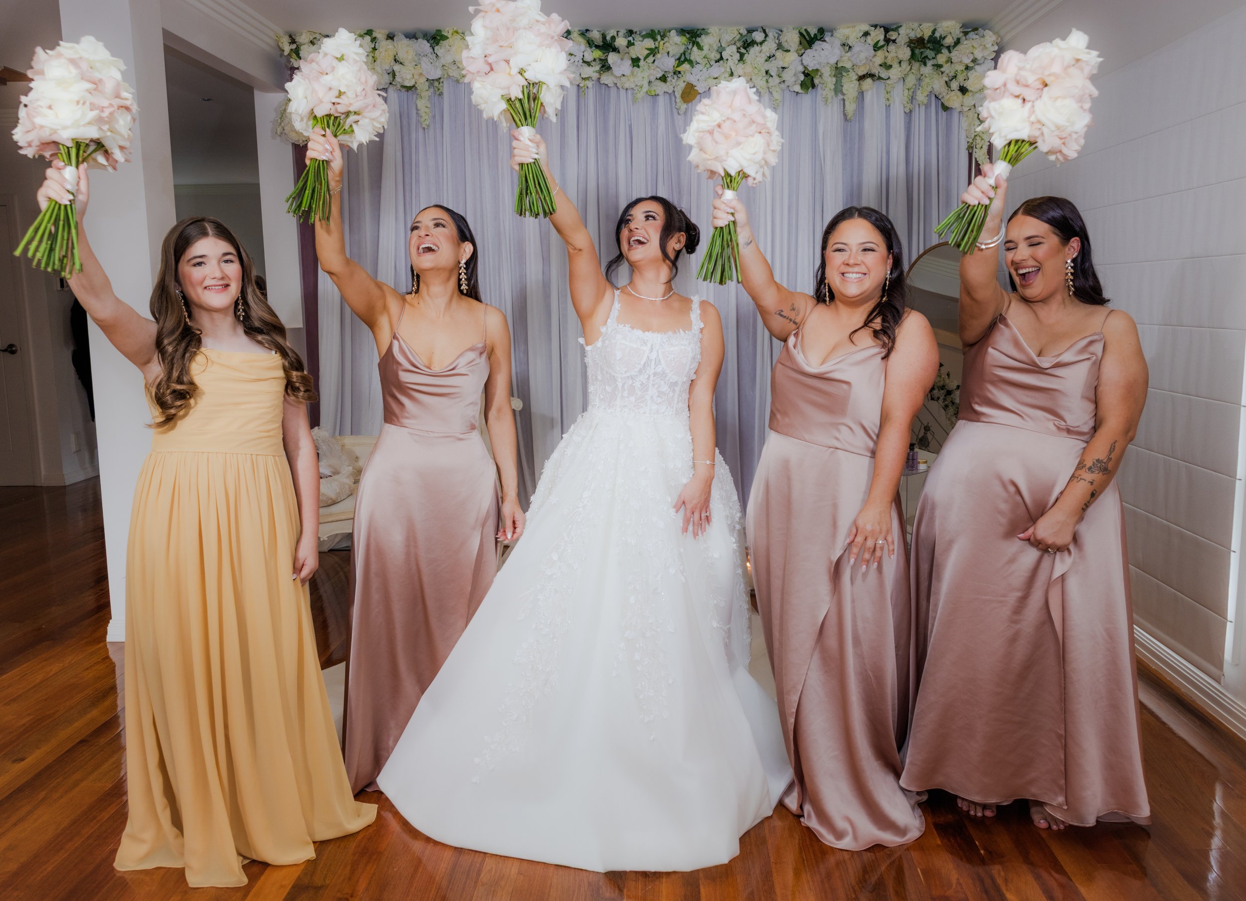 A bride and four bridesmaids celebrating with bouquets in hand at a wedding. The bride is in the center wearing a white lace wedding dress, with bridesmaids in satin dresses around her. All are smiling and cheering, standing in front of a floral back