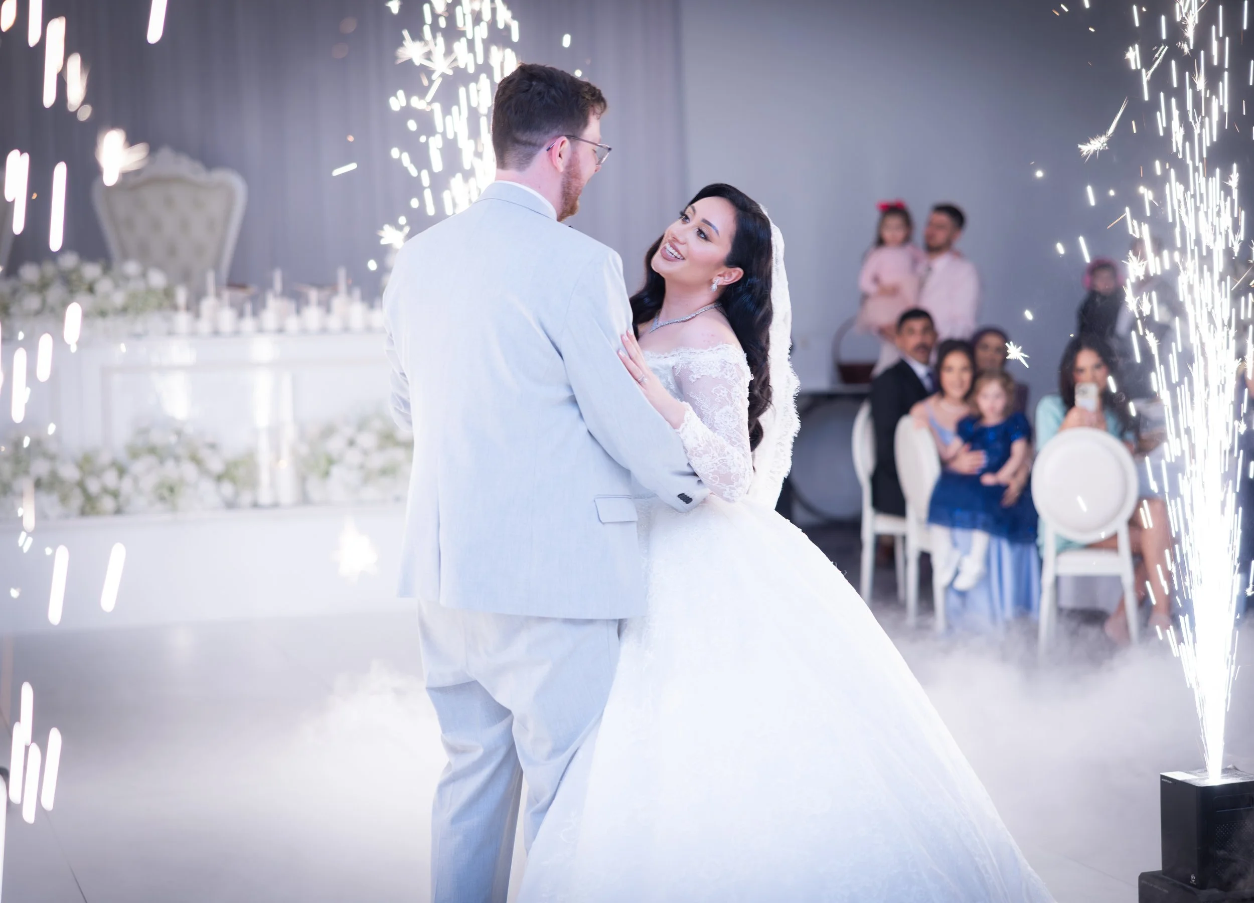 A bride and groom dancing at their wedding reception, with sparklers in the background and wedding guests seated watching. The bride is smiling at the groom, who is wearing a light-colored suit.