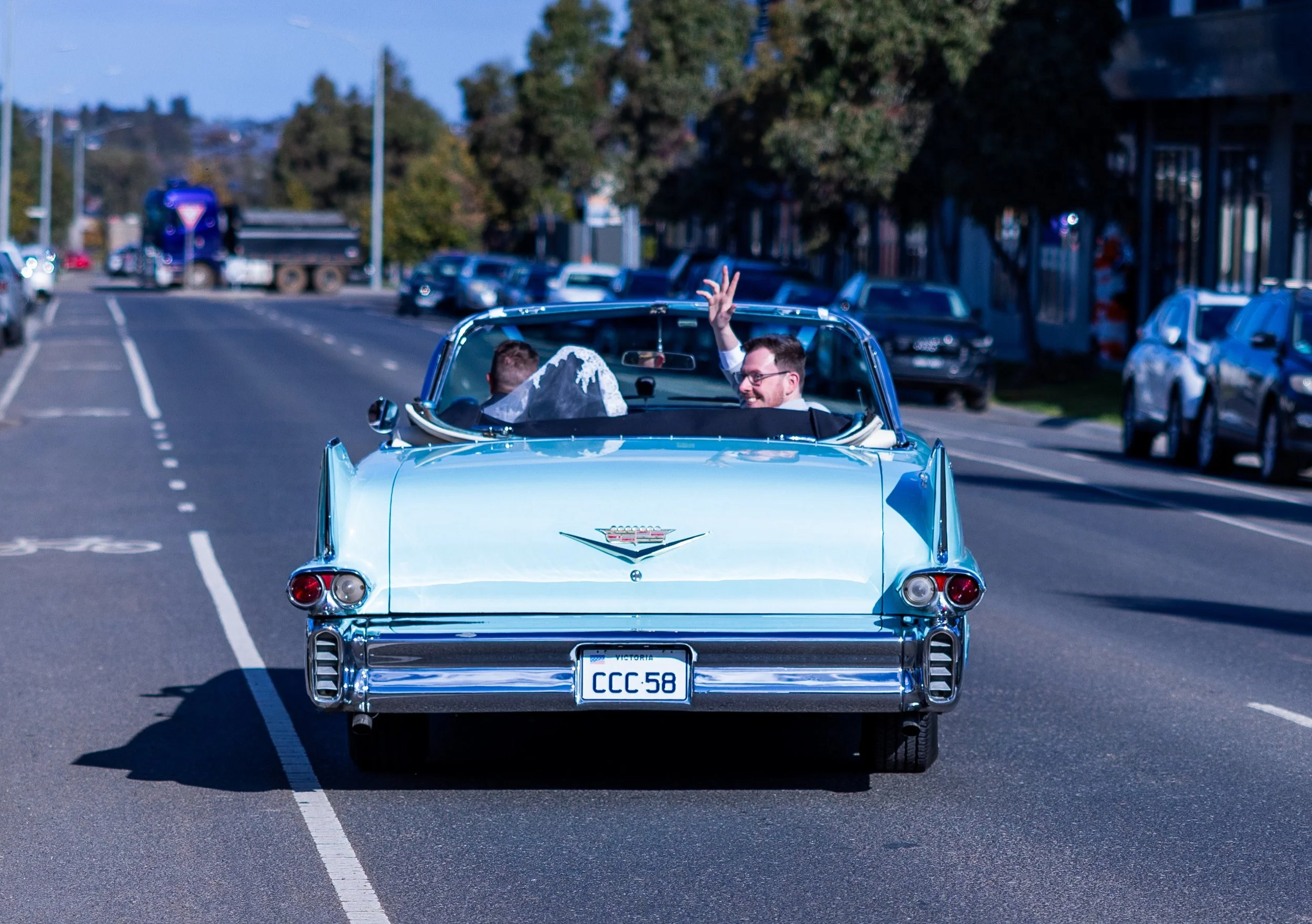 A vintage blue convertible car with a mountain emblem on the back, driven by a smiling man with glasses who is waving, and a woman in a white dress with an umbrella, driving on a city street.