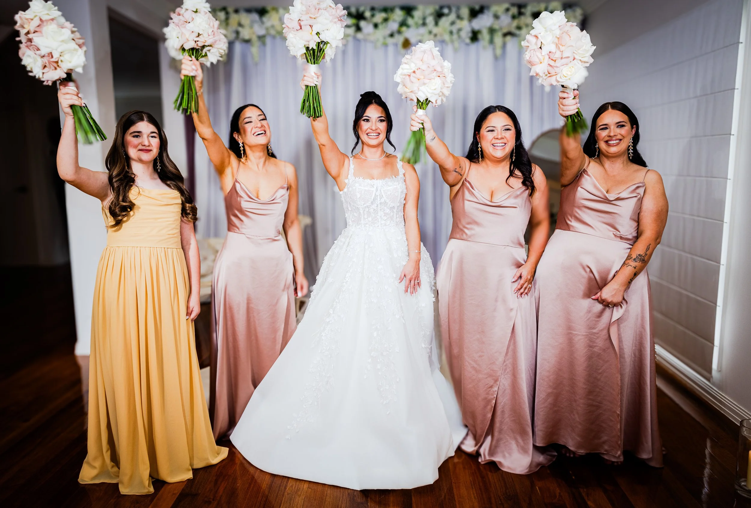 Bride in a white wedding gown holding a bouquet of pink flowers, surrounded by four bridesmaids in blush pink satin dresses and one in a yellow dress, all smiling and raising bouquets in celebration.