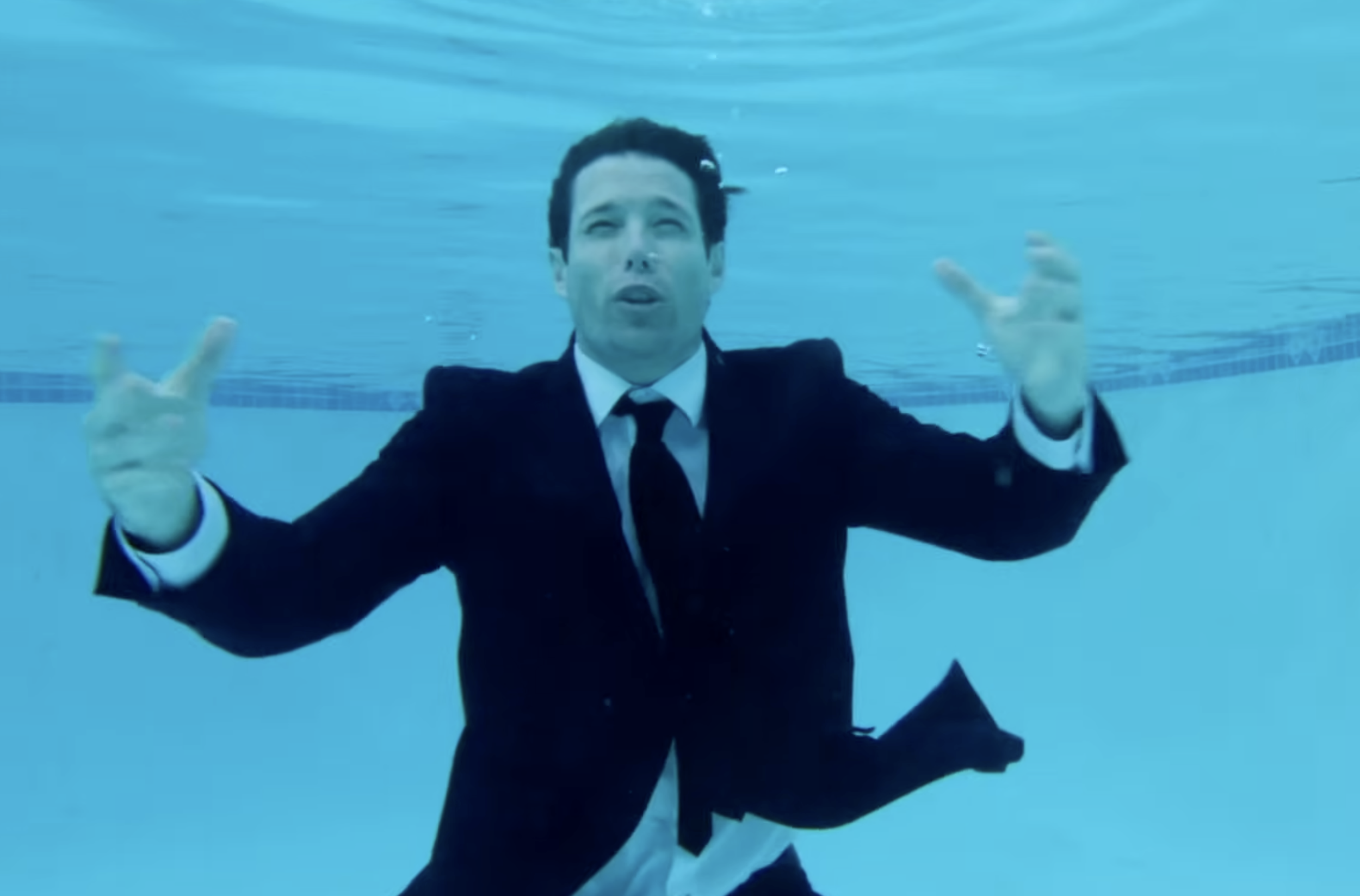 A man in a black suit and tie is underwater in a swimming pool, making hand gestures.
