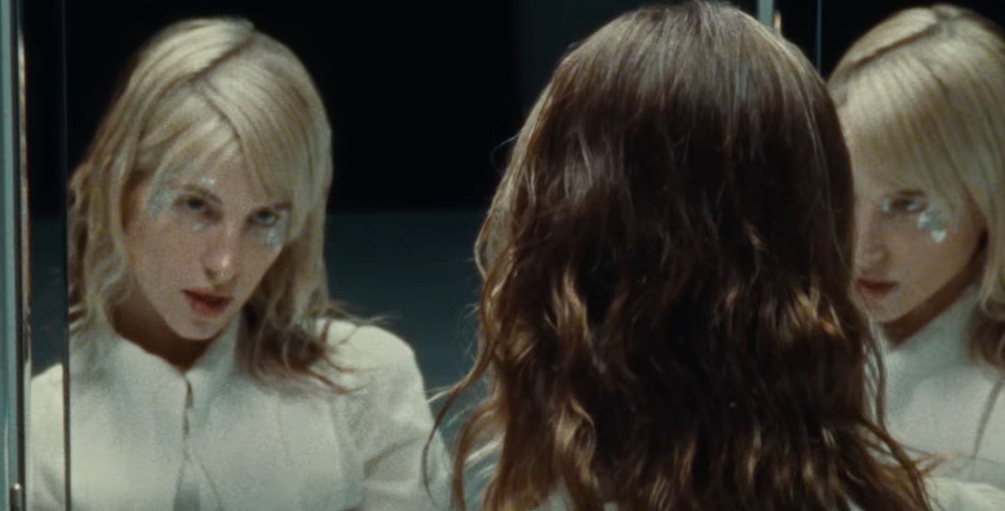 A woman with wavy brown hair looking into a mirror and seeing her reflection, which shows her with blonde hair and makeup with star-shaped face paint.