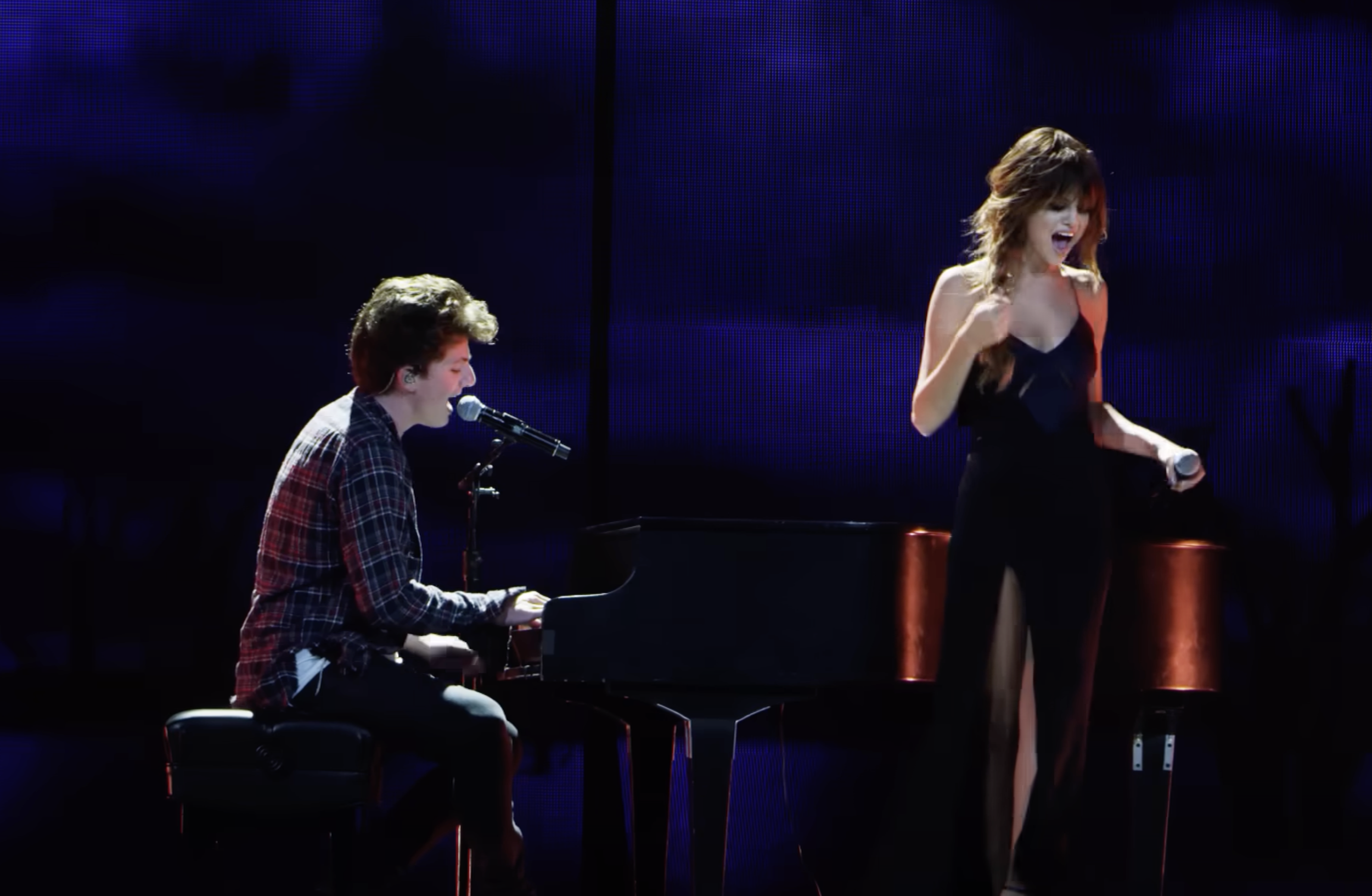 A young man playing a black grand piano and singing into a microphone on a dark stage, while a young woman in a black dress with a high slit sings into a microphone nearby.