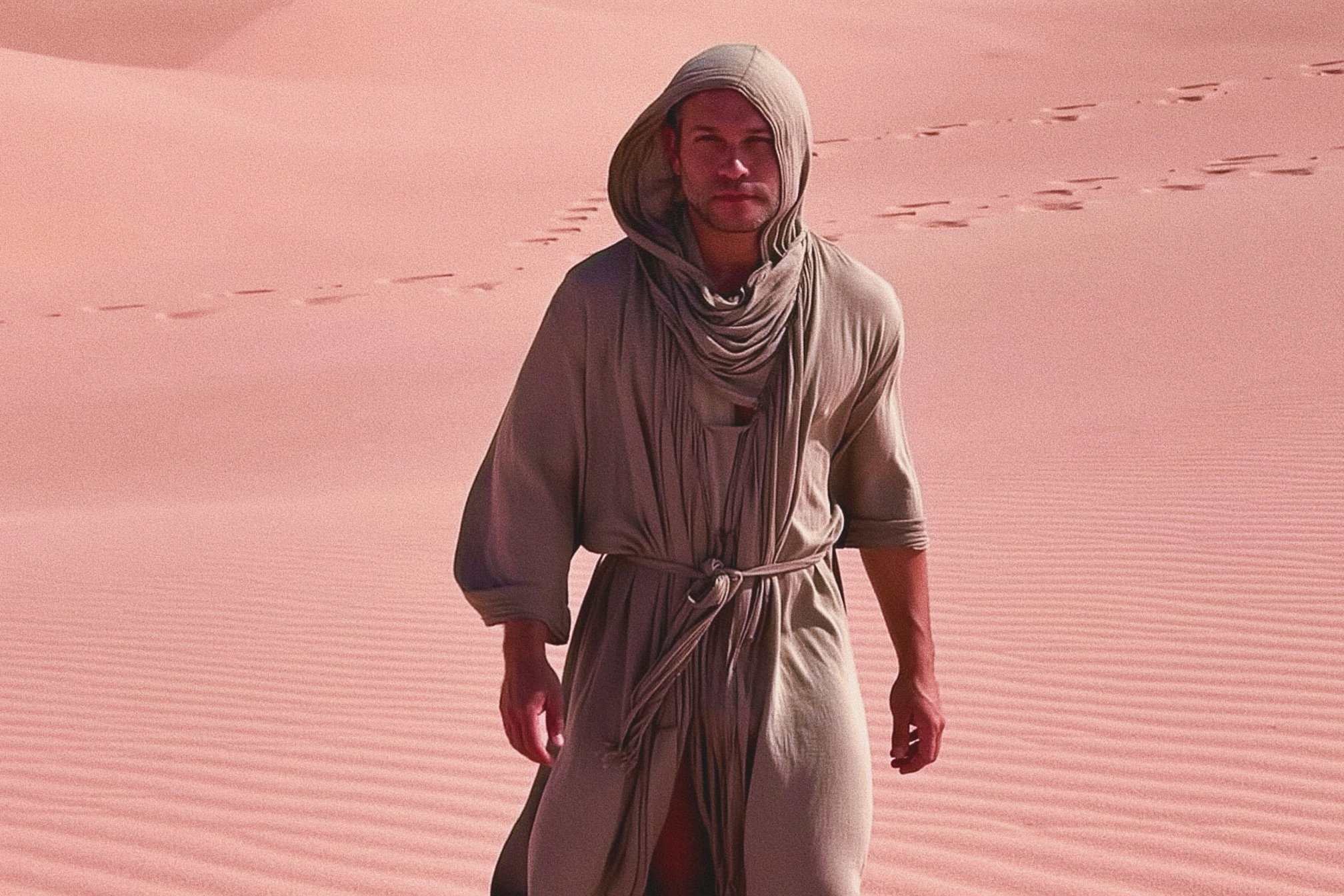 A man dressed in beige robes and a head covering standing on a desert with pink sand, footprints in the background.