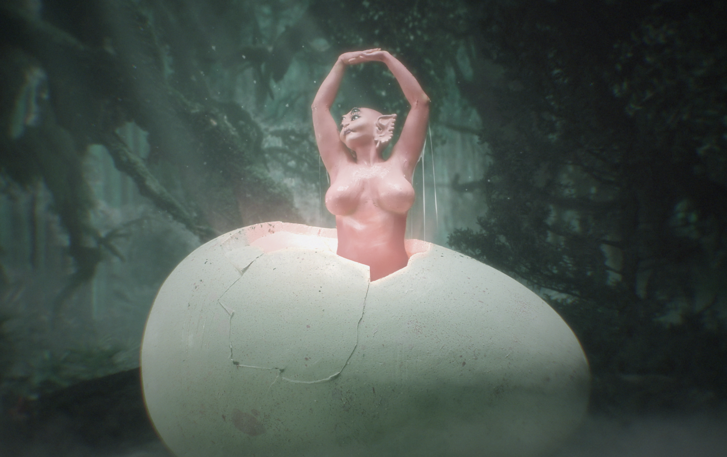 A fantasy sculpture of a humanoid cat with human female torso emerging from a cracked eggshell in a forest setting.