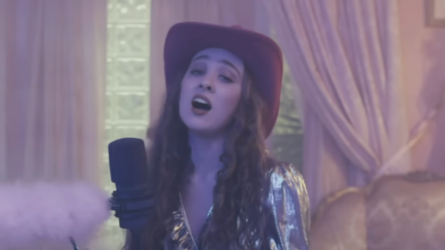 A woman with long, curly hair wearing a large-brimmed hat and a shiny, metallic jacket, singing into a microphone in a room with purple curtains and golden decor.