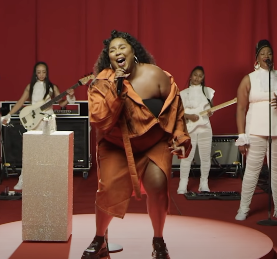 A woman singing passionately on stage with a band of women playing guitars and keyboards against a red background.