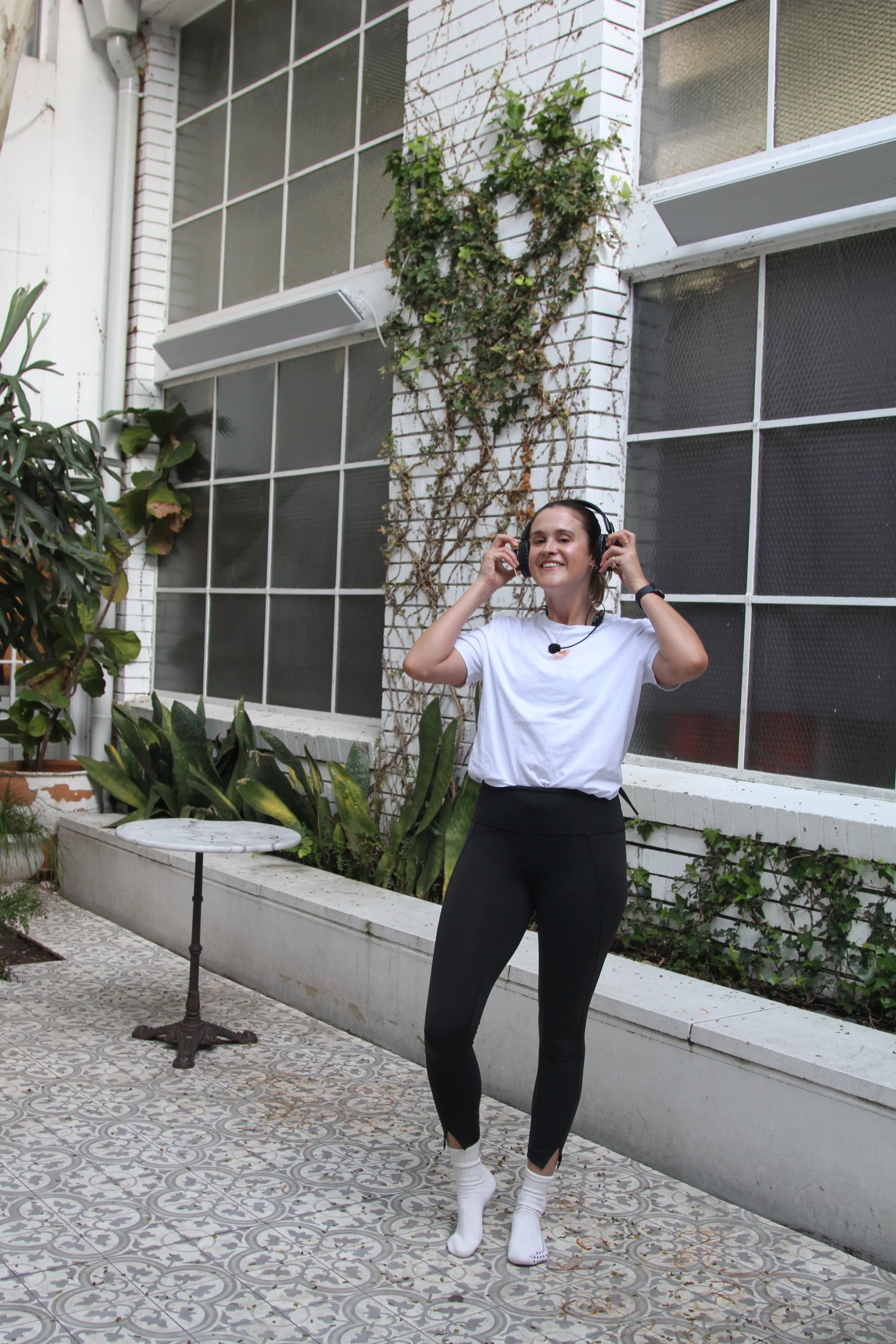 A woman smiling and enjoying listening to music with headphones in an outdoor patio area. She is wearing a white t-shirt, black leggings, and white socks. There are plants and a patterned tiled floor around her, with a building wall covered in ivy in the background.