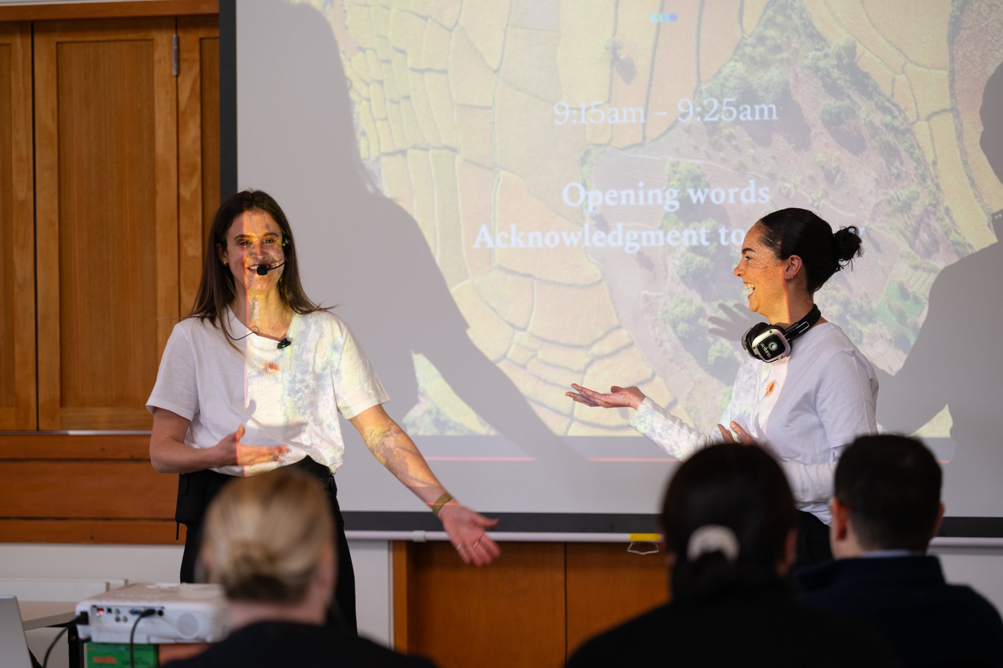 Two women giving a presentation in front of a projection screen in a classroom. One woman is smiling and gesturing with her hands, while the other woman is smiling and has headphones around her neck. There are audience members in the foreground.