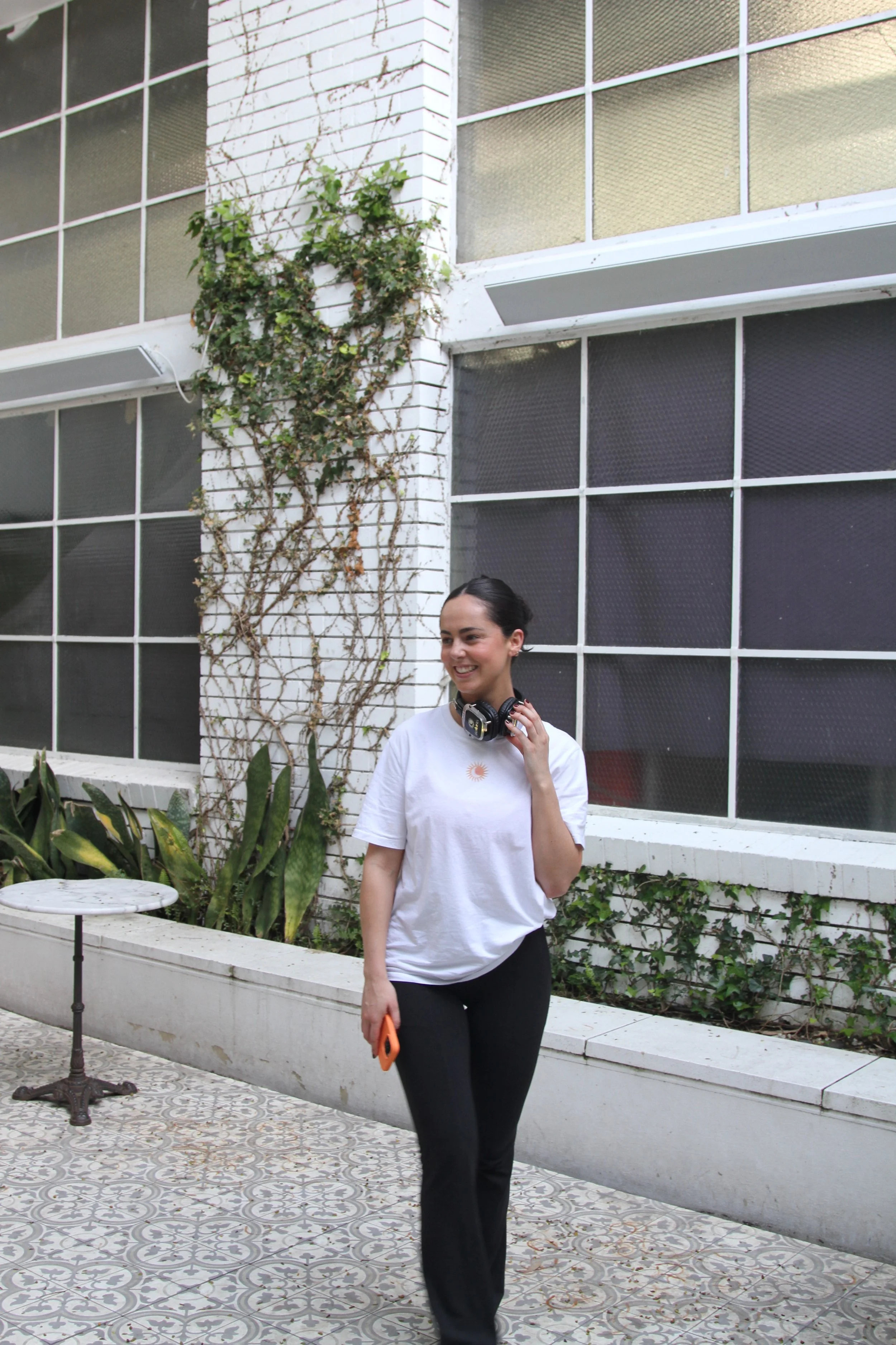 Woman in white t-shirt and black pants walking outdoors near a building with large windows and vines on the wall, holding a phone and a pair of headphones.