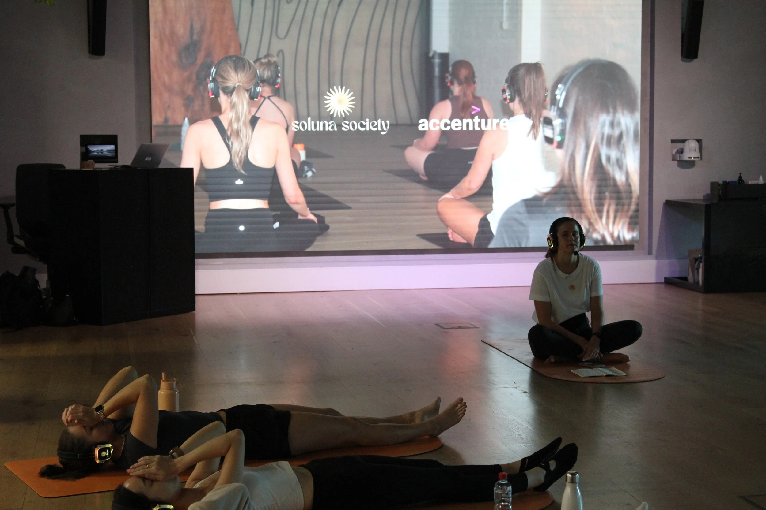 People participating in a virtual fitness or meditation class, wearing headphones, lying on yoga mats, with a large screen displaying a yoga session with women sitting cross-legged.