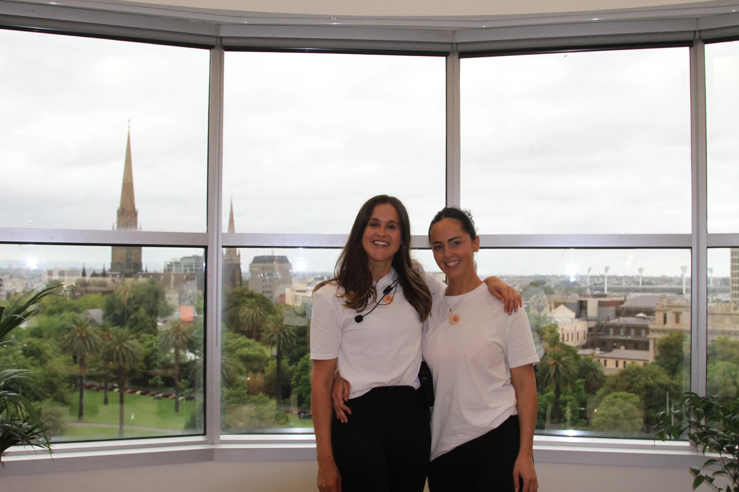 Two women smiling and standing together in front of a large window with a cityscape and church spires in the background.