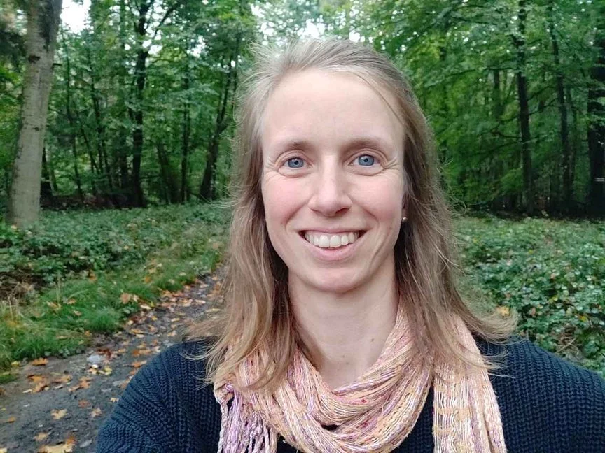 Frau lächelt in einem Waldweg, umgeben von grünen Bäumen.