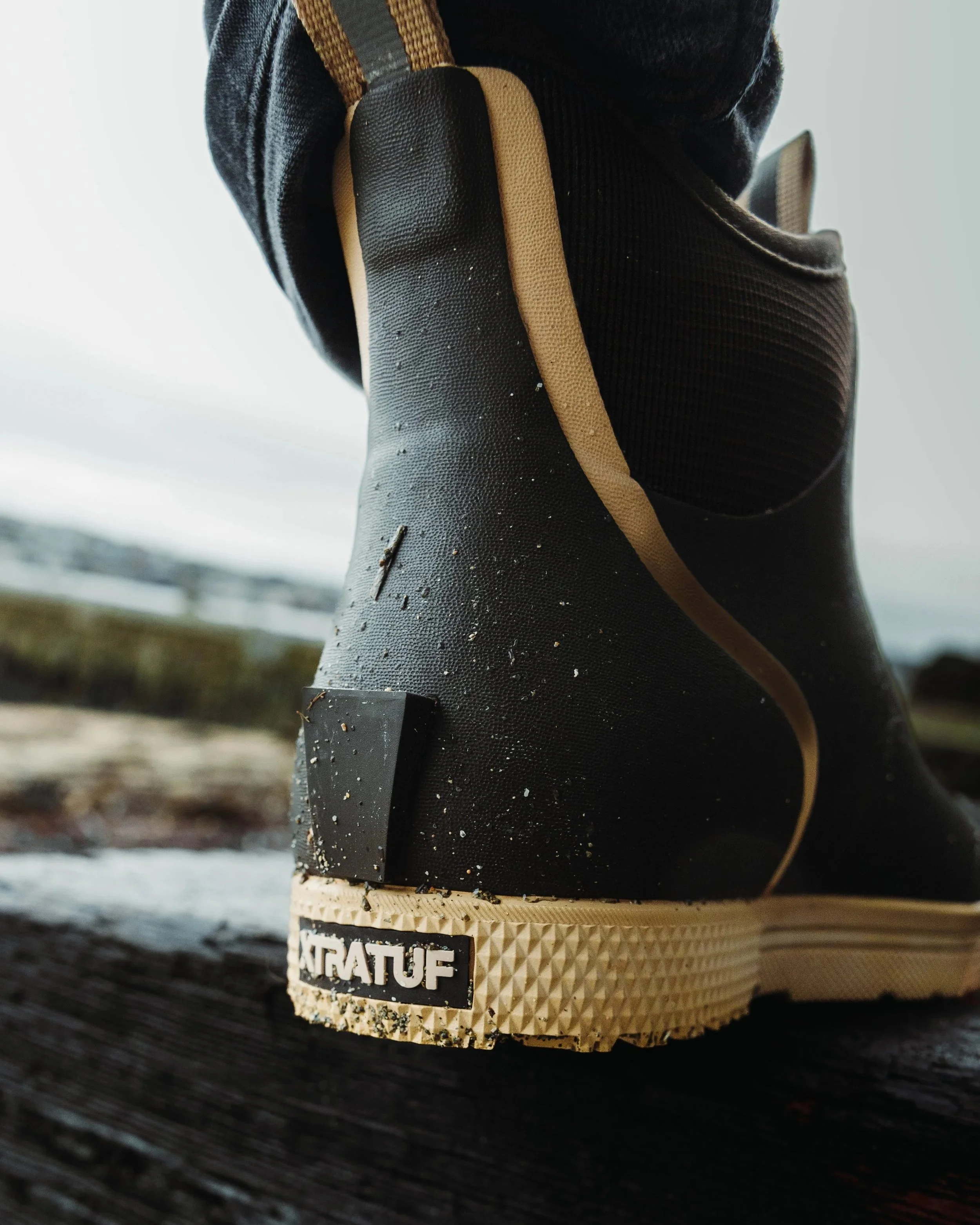 Close-up of a black rubber outdoor boot with beige sole, sitting on a wooden surface, with a blurred outdoor background.