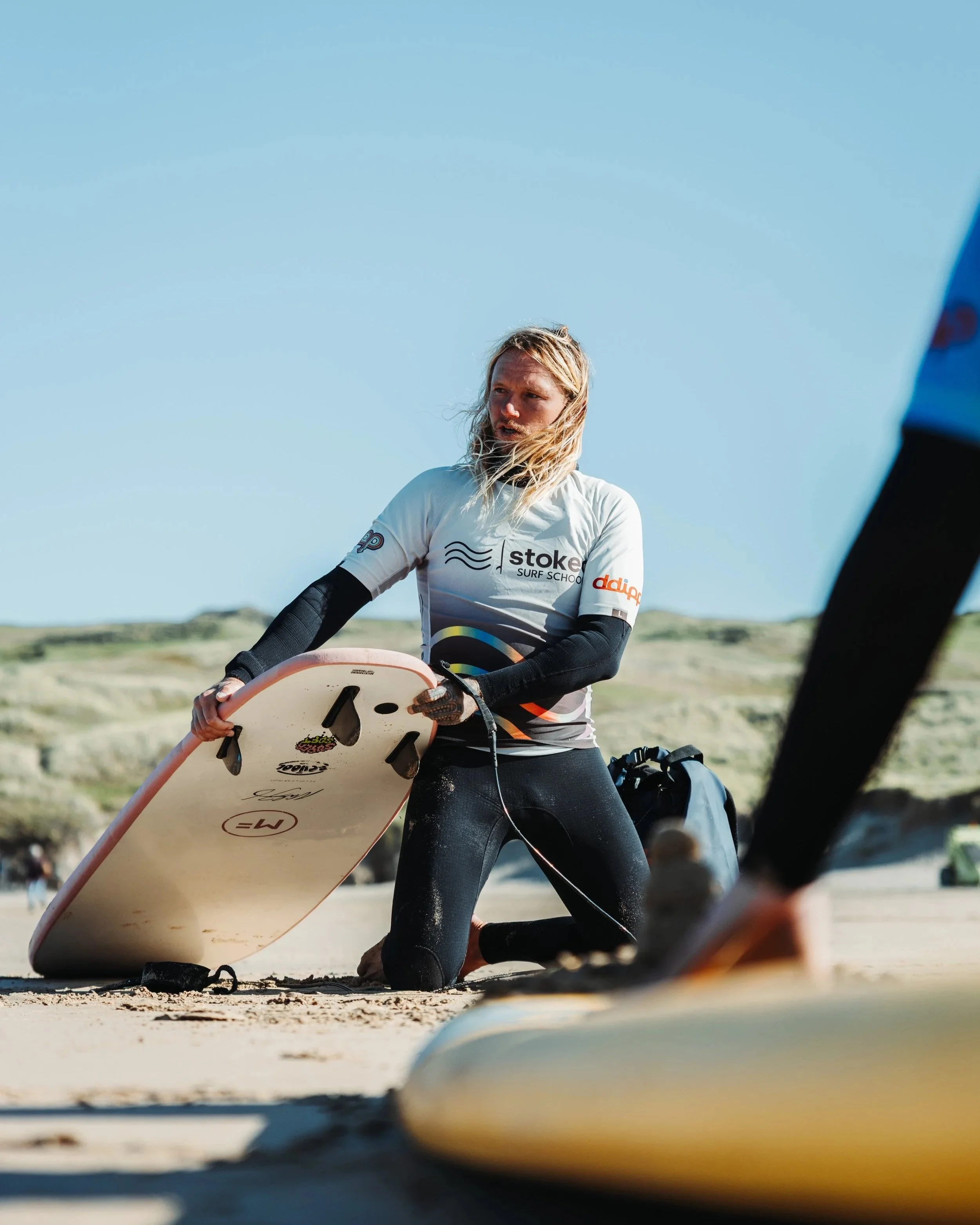 A woman kneeling on a sandy beach, holding a surfboard, with sandy dunes and sky in the background.