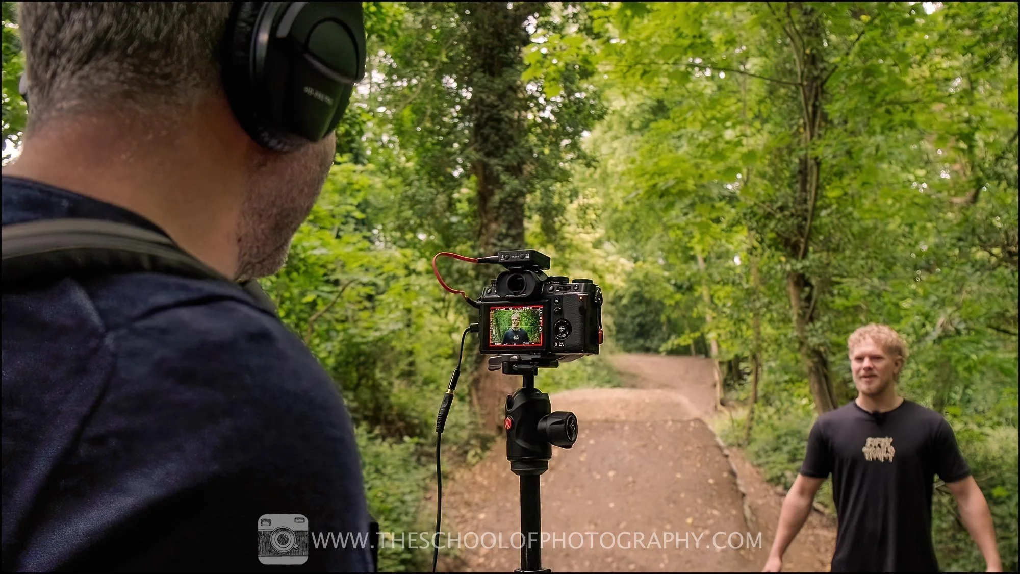 Photographer filming a person speaking on a forest trail using a mirrorless camera mounted on a tripod, illustrating the practice of capturing subjects and storytelling in photography.