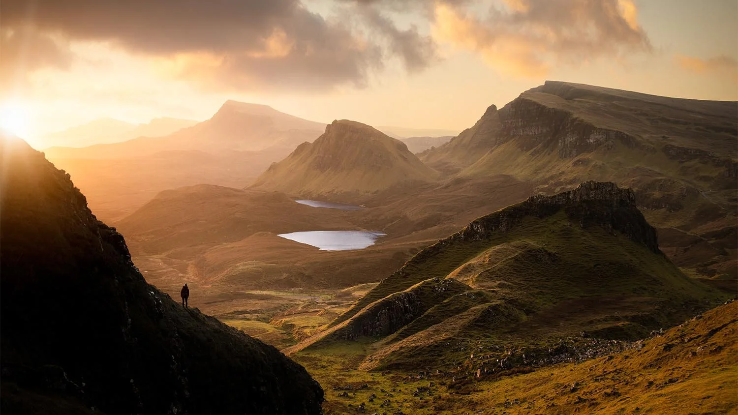 Mountain landscape at sunrise with a lone hiker overlooking valleys and lakes during golden hour.