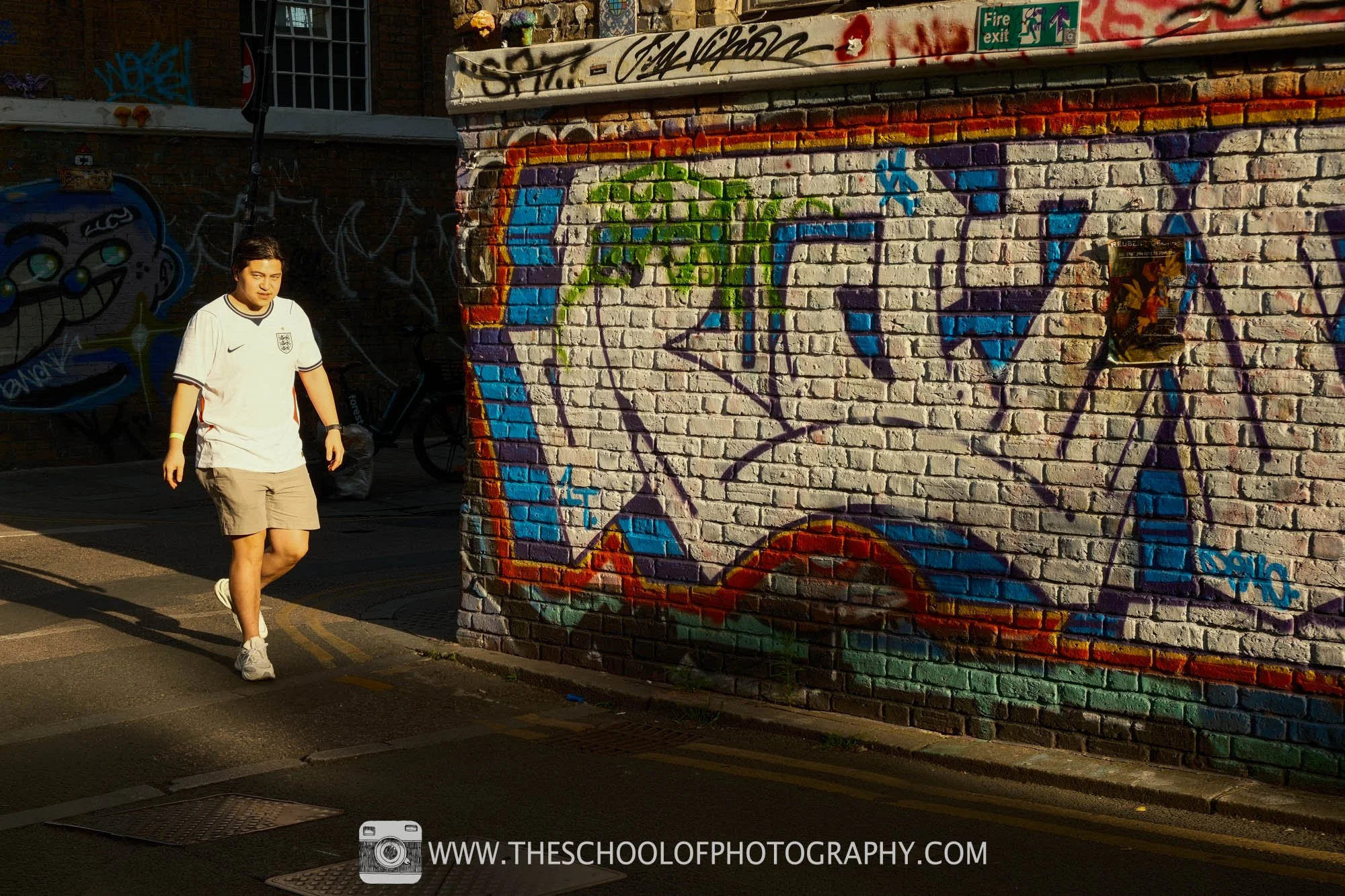 A man in a white shirt walks through warm sunlight beside a colourful graffiti-covered brick wall.
