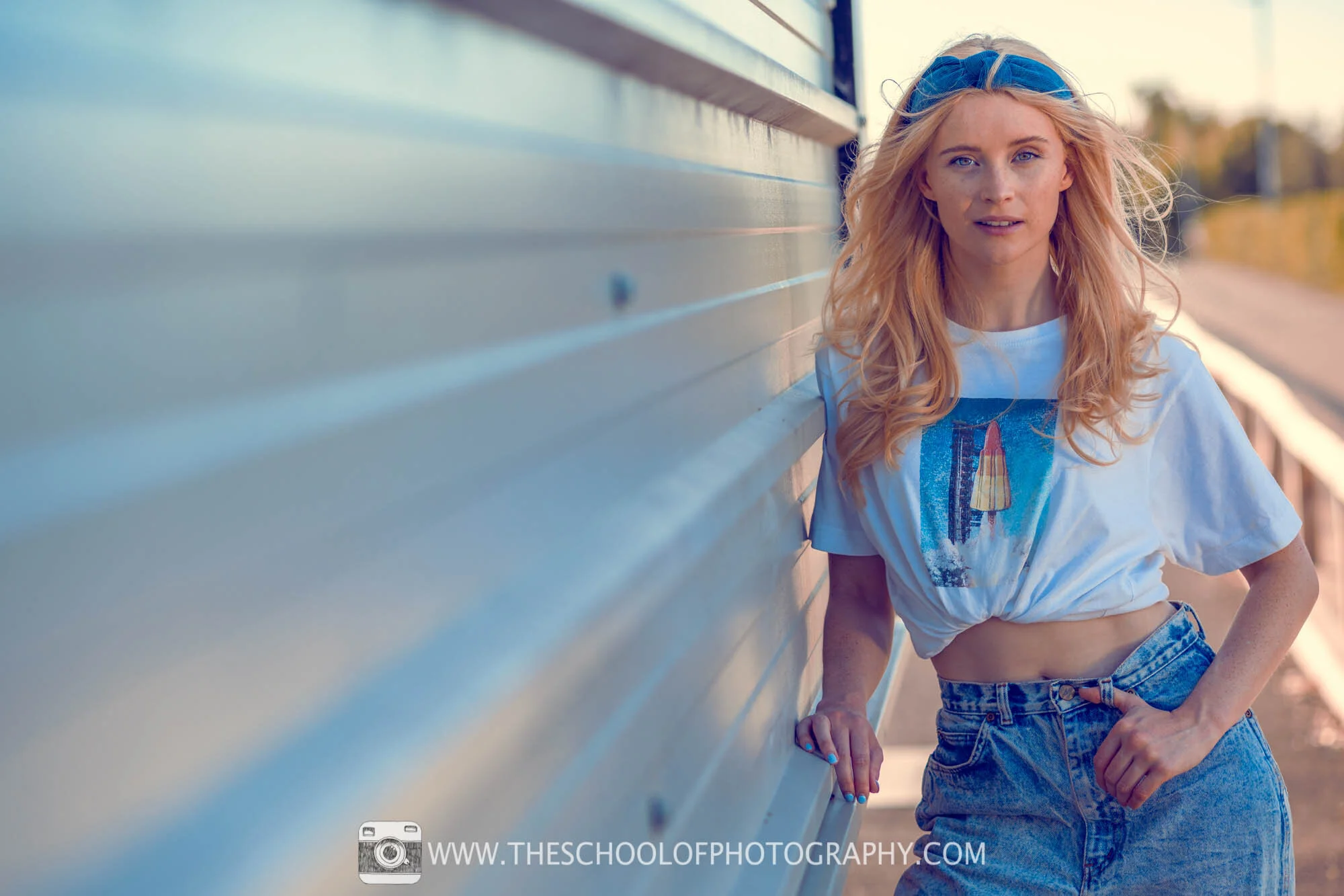 Outdoor portrait of a blonde woman leaning against a wall, wearing a white T-shirt and denim shorts.
