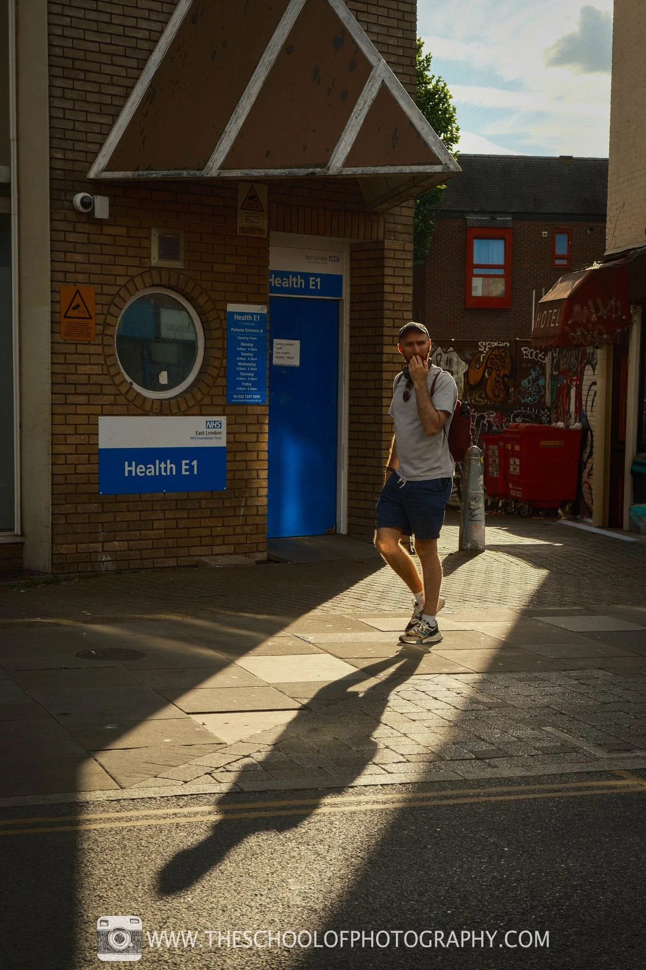 A man walks through a shaft of warm sunlight on a city street, casting a long shadow across the pavement.