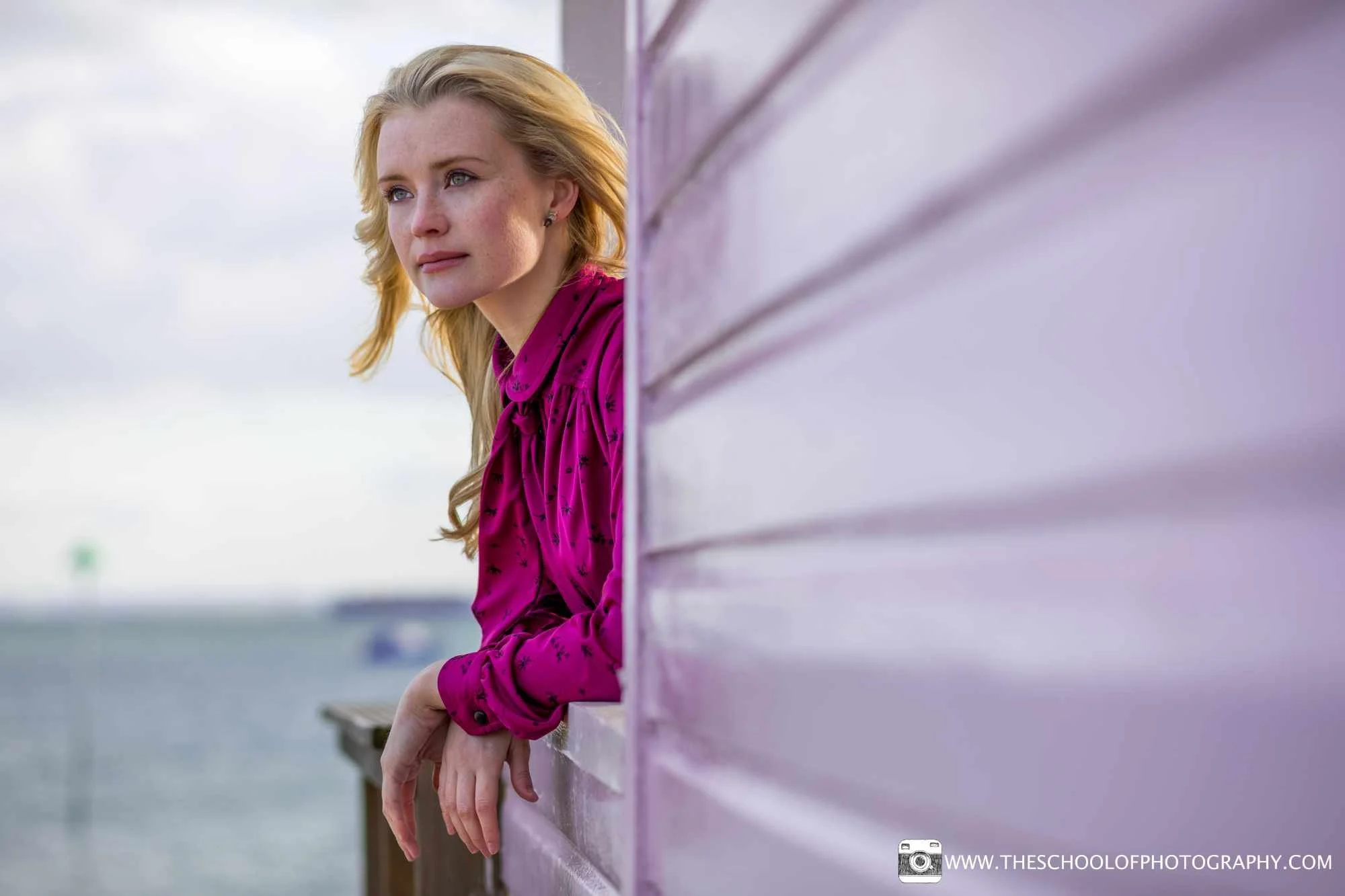 Portrait of a woman in a bright pink blouse leaning against a pink beach hut beside the sea.