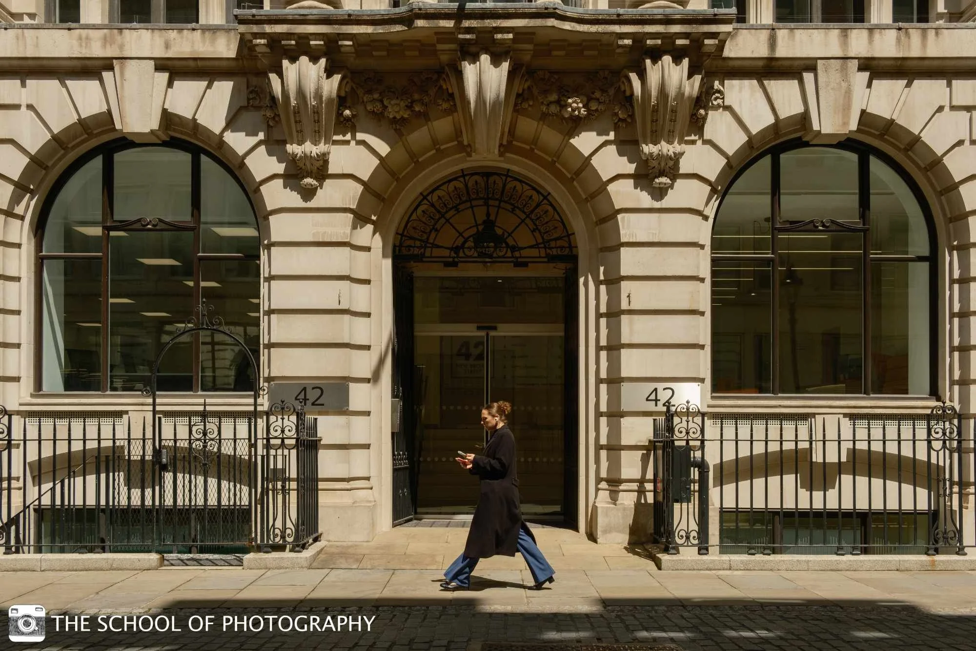 Colour street photography image of a woman walking past an ornate London building in strong sunlight, using architectural symmetry, shadows and warm tones.