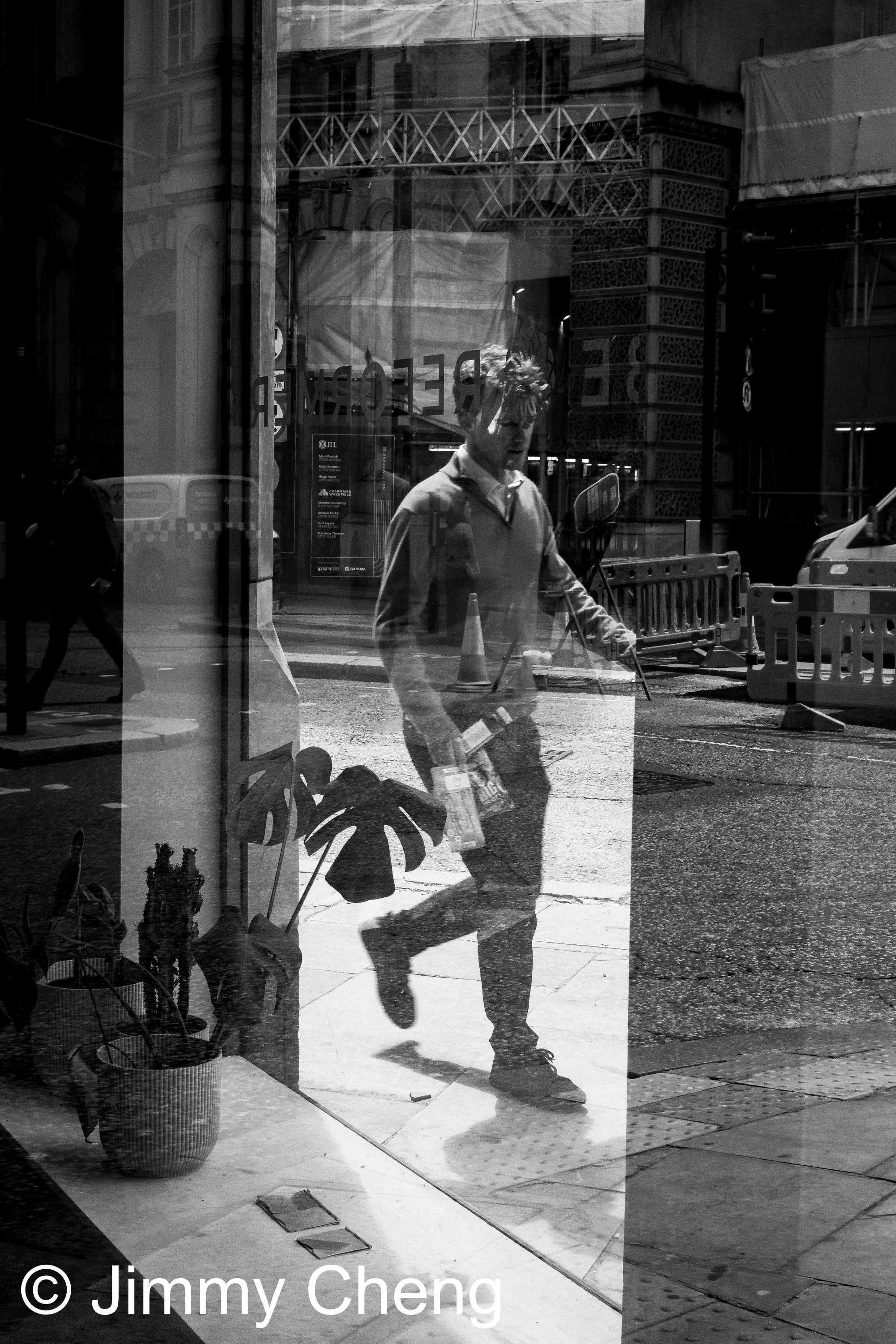 Black and white street photography showing a man walking past a reflective shop window, layering his figure with reflections, architecture and strong light and shadow.