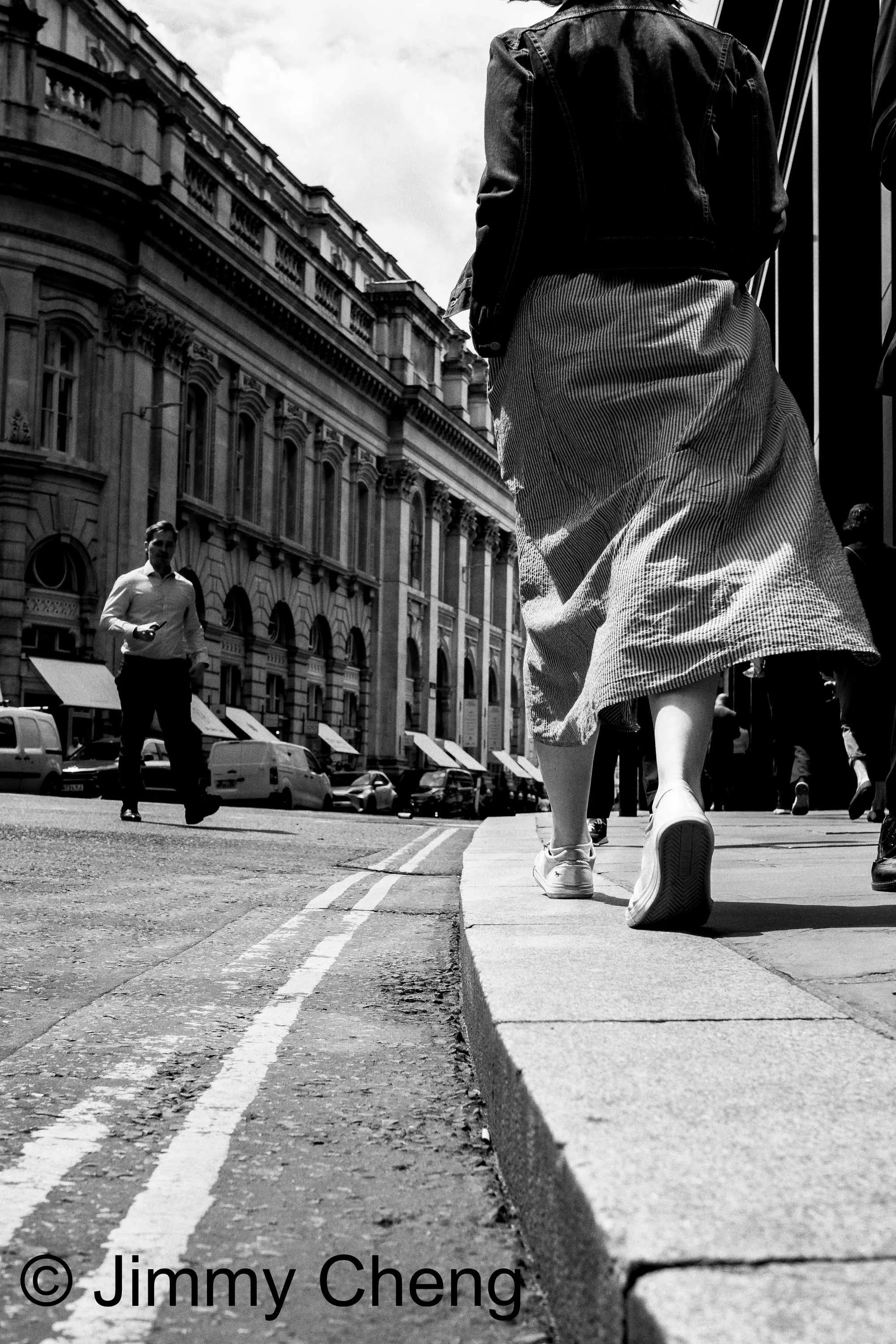 Black and white street photography taken from a low angle, showing pedestrians walking through a city street with strong leading lines, harsh light and deep shadows.