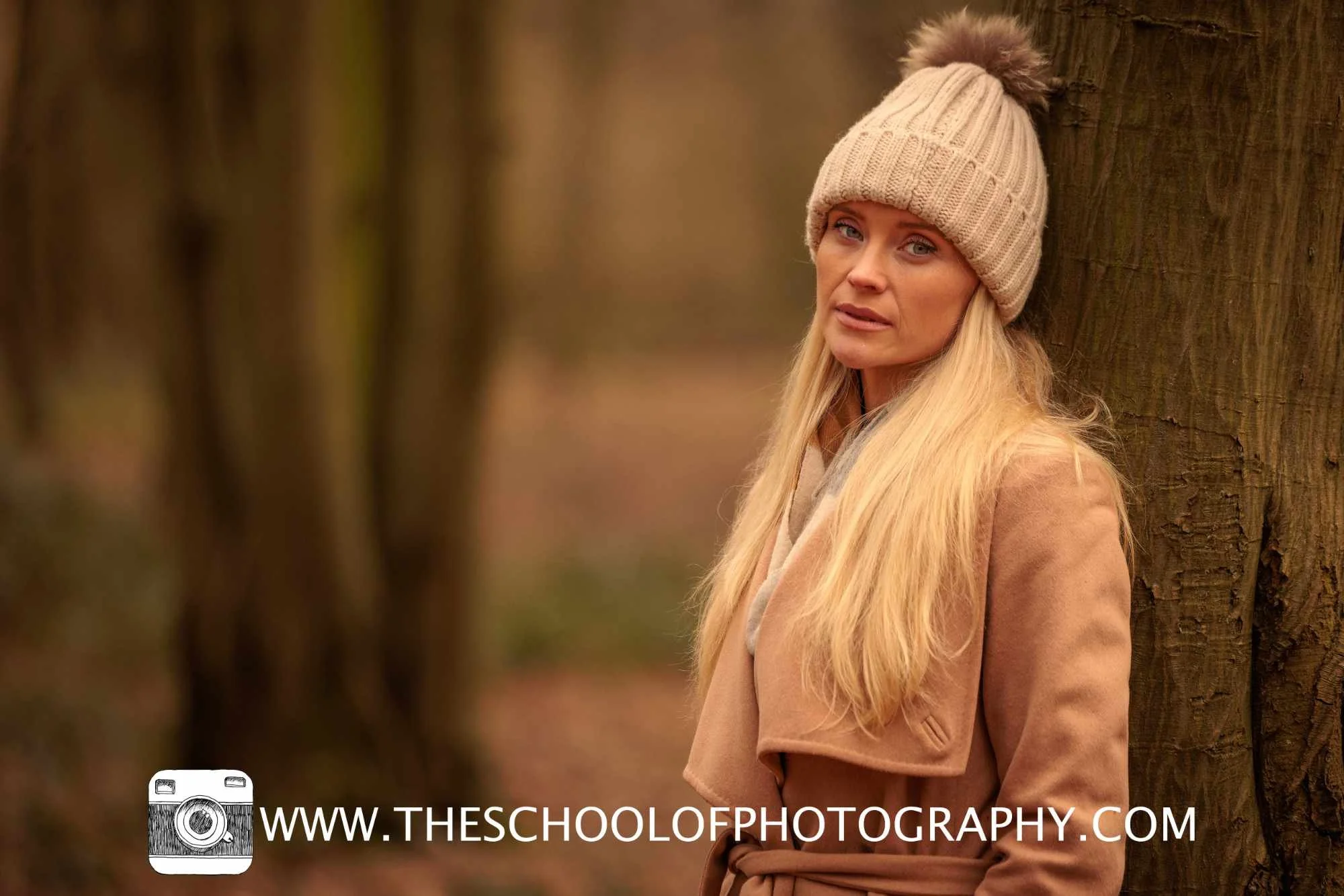 A woman in a knit beanie and tan coat stands beside a tree in a softly blurred woodland setting, with her face and eyes sharply focused against the out-of-focus background.