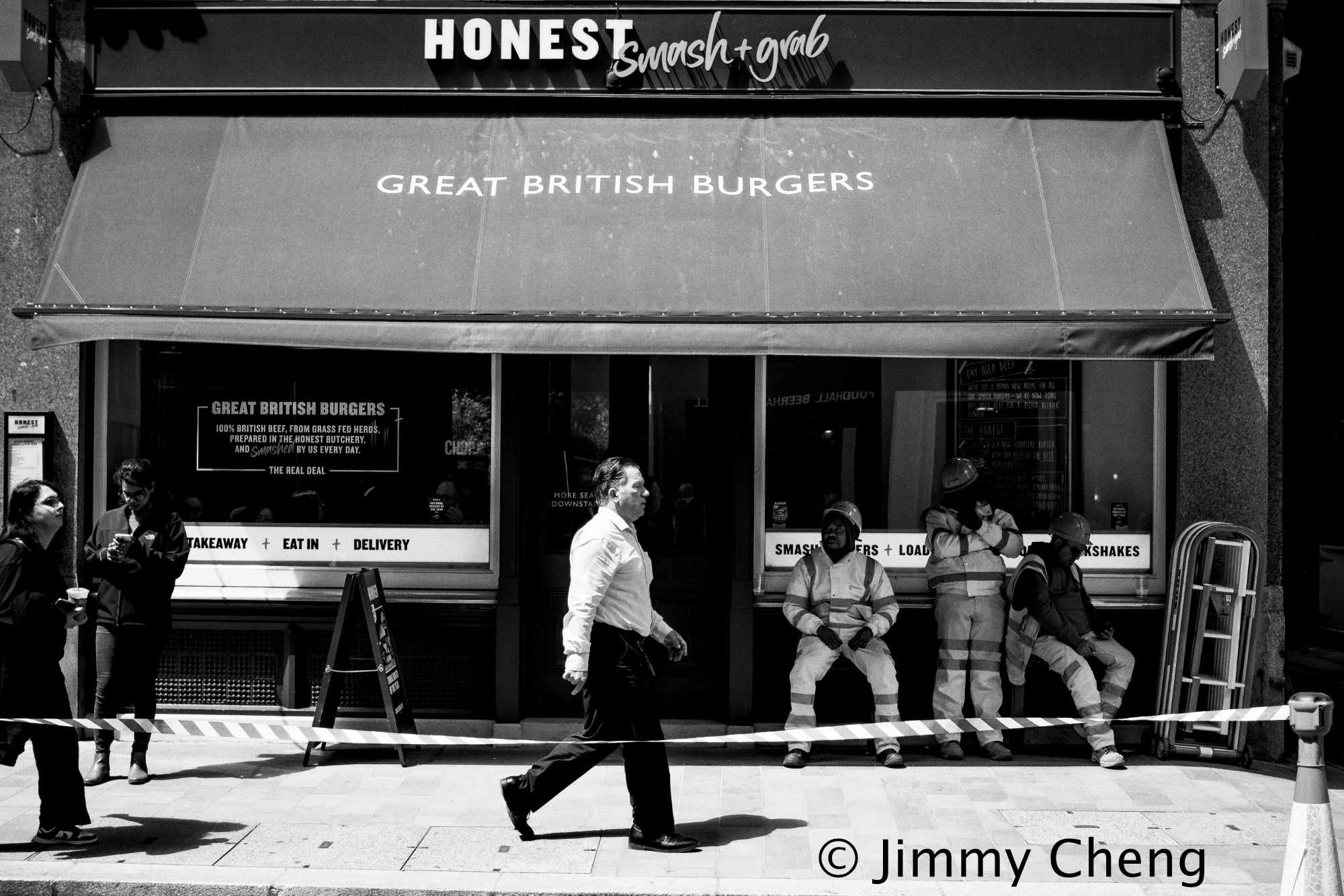 Black and white street photography showing pedestrians and construction workers outside a London burger restaurant, with a man walking through the centre of the scene.