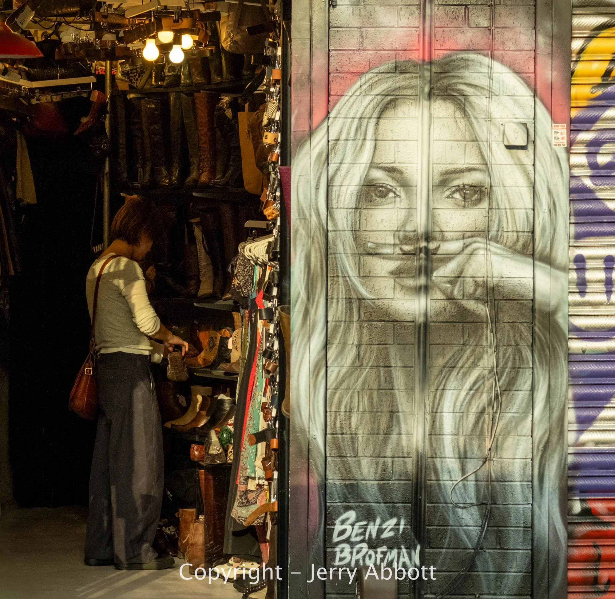 A shopper browses boots inside a market stall beside a large monochrome portrait painted on a brick wall.