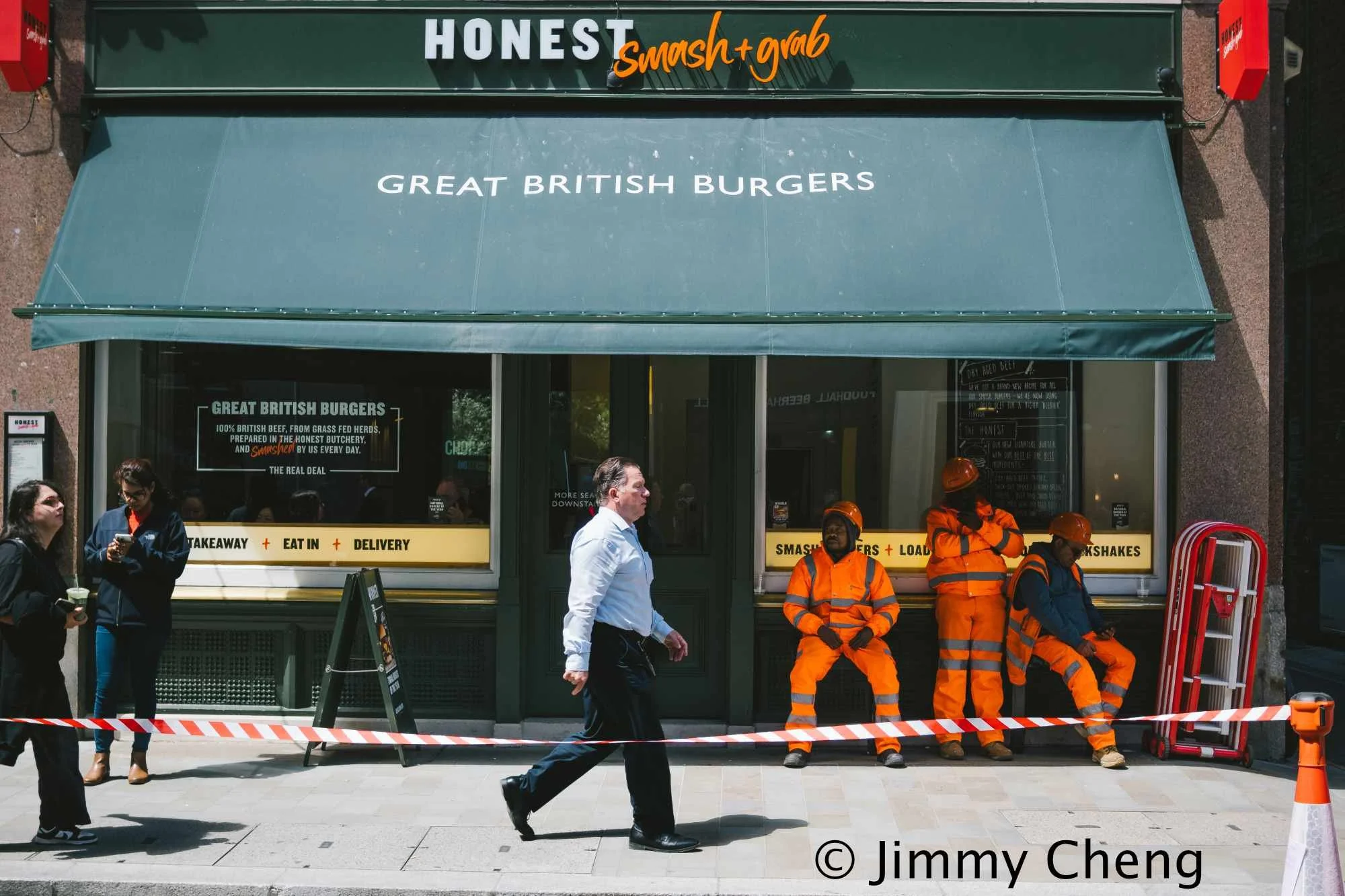 Colour street photography showing pedestrians and construction workers in bright orange clothing outside a London burger restaurant, with a man walking through the centre.