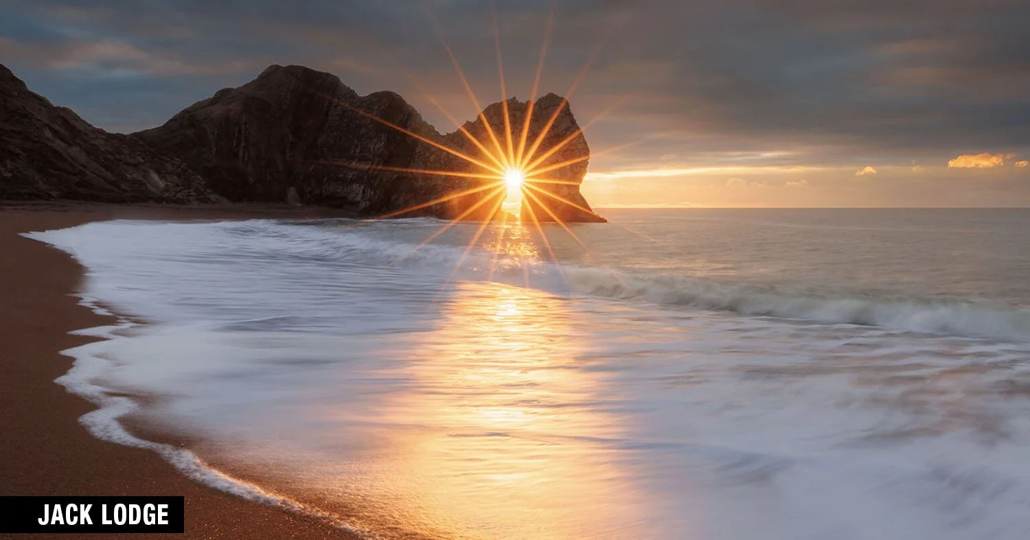 Durdle Door sea arch at sunrise with sunburst and waves on the beach.