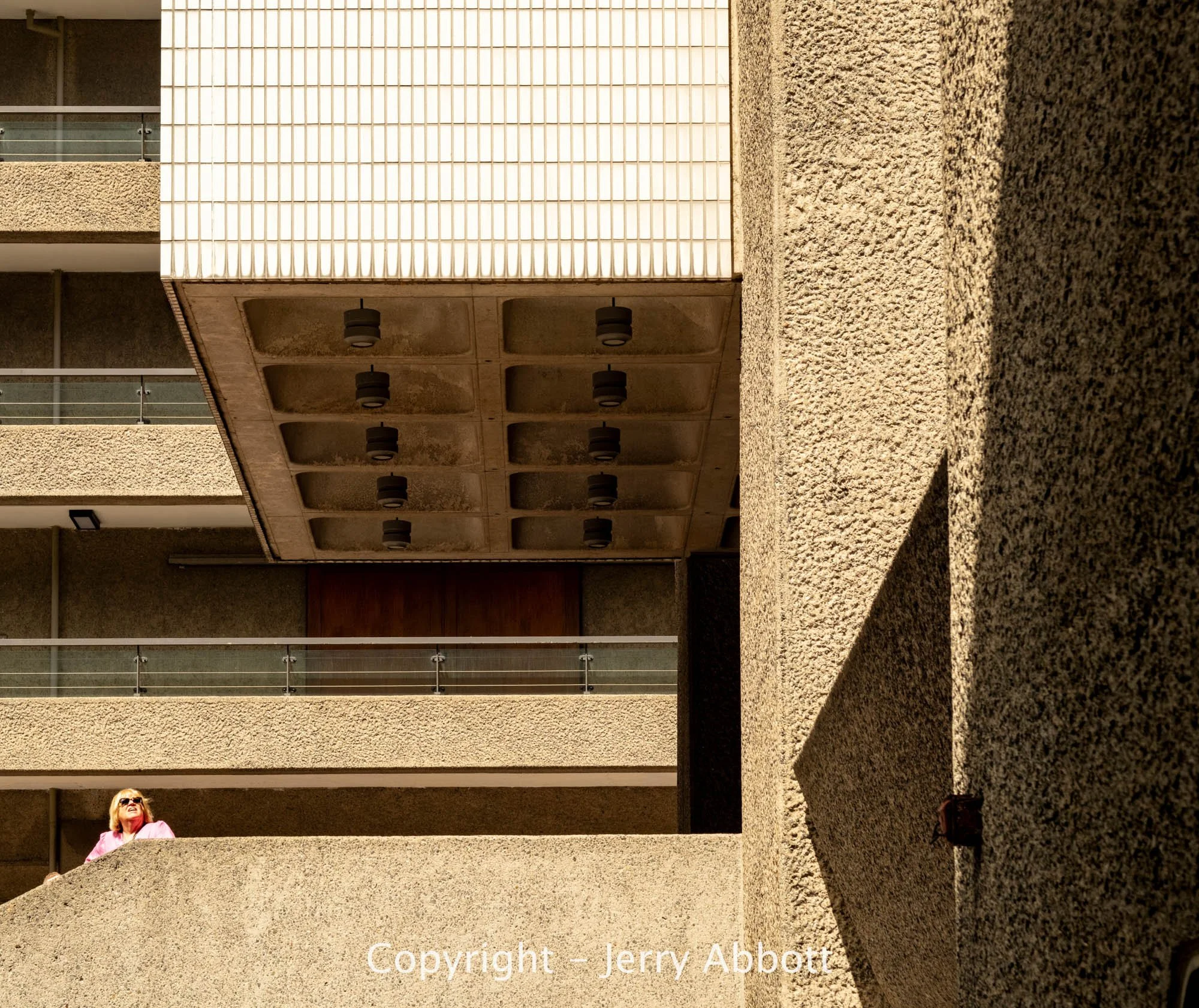 A woman in pink stands within the geometric concrete forms of Brutalist architecture, illuminated by warm sunlight and strong shadows.
