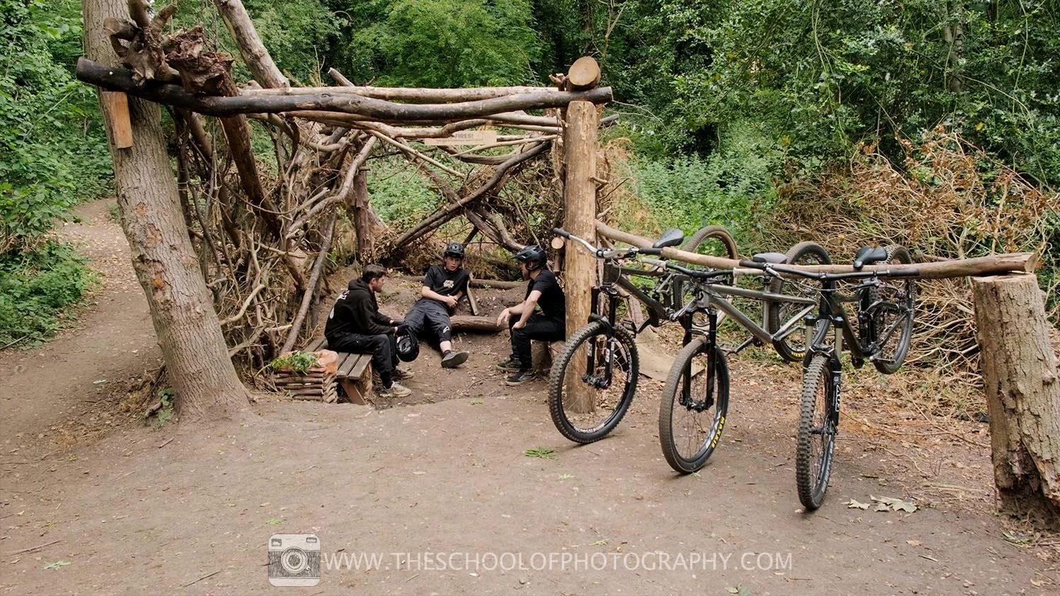 Wide shot of three cyclists resting in a wooded trail area with their mountain bikes nearby, showing the full environment and context.