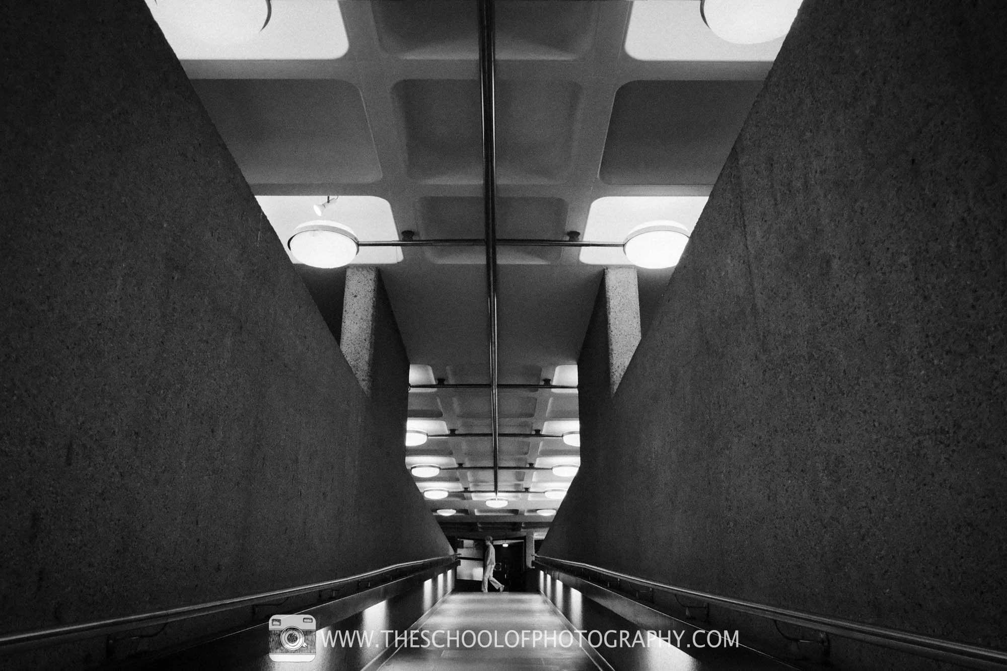 Black-and-white view down a dramatic Brutalist walkway, with repeating ceiling lights and a lone figure in the distance.