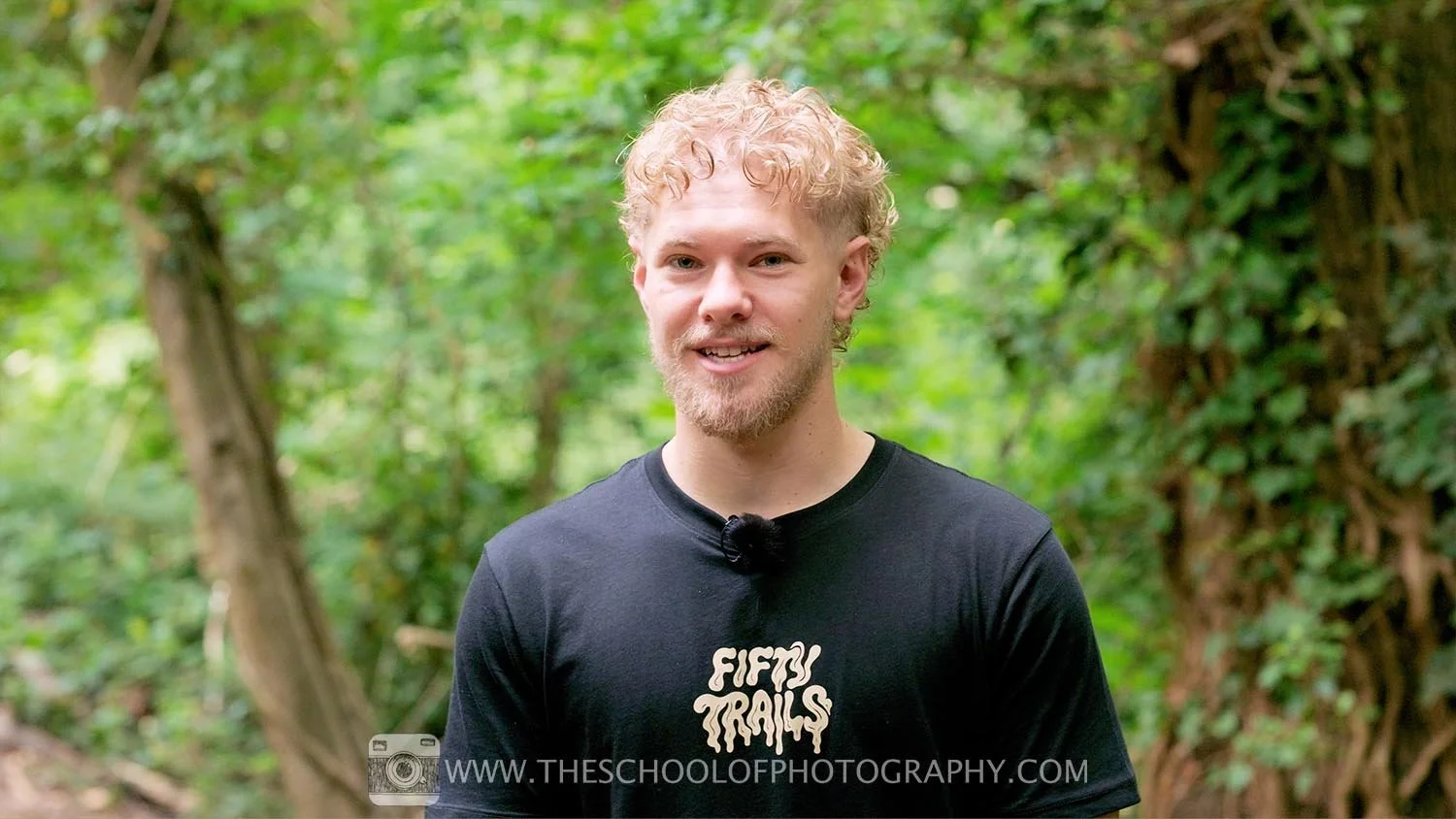 Medium shot of a man speaking to camera outdoors in a wooded area, with a blurred natural background.