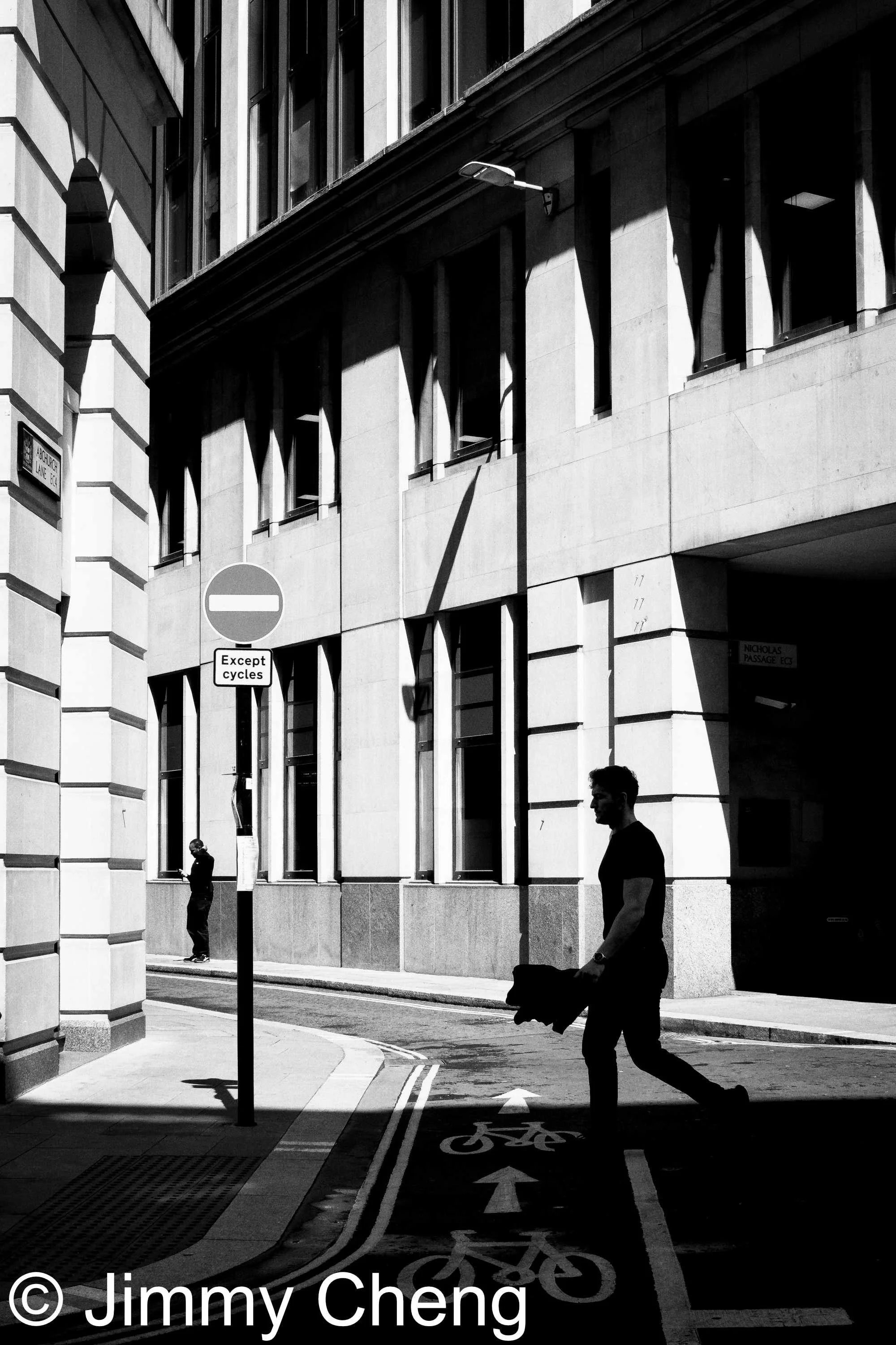 Black and white London street photography using harsh sunlight, deep shadows and strong architectural lines, with a silhouetted pedestrian crossing the street.