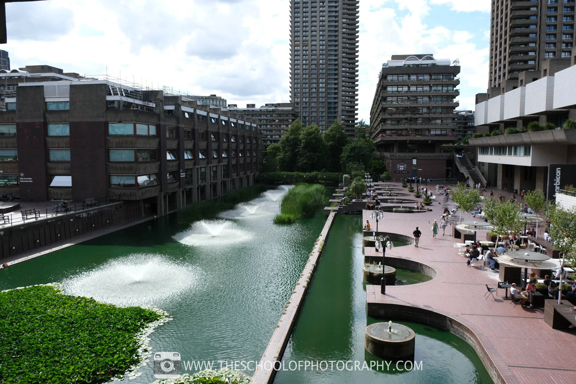 Elevated view of the Barbican in London, with fountains across a green lake, a busy waterside terrace and surrounding Brutalist buildings.
