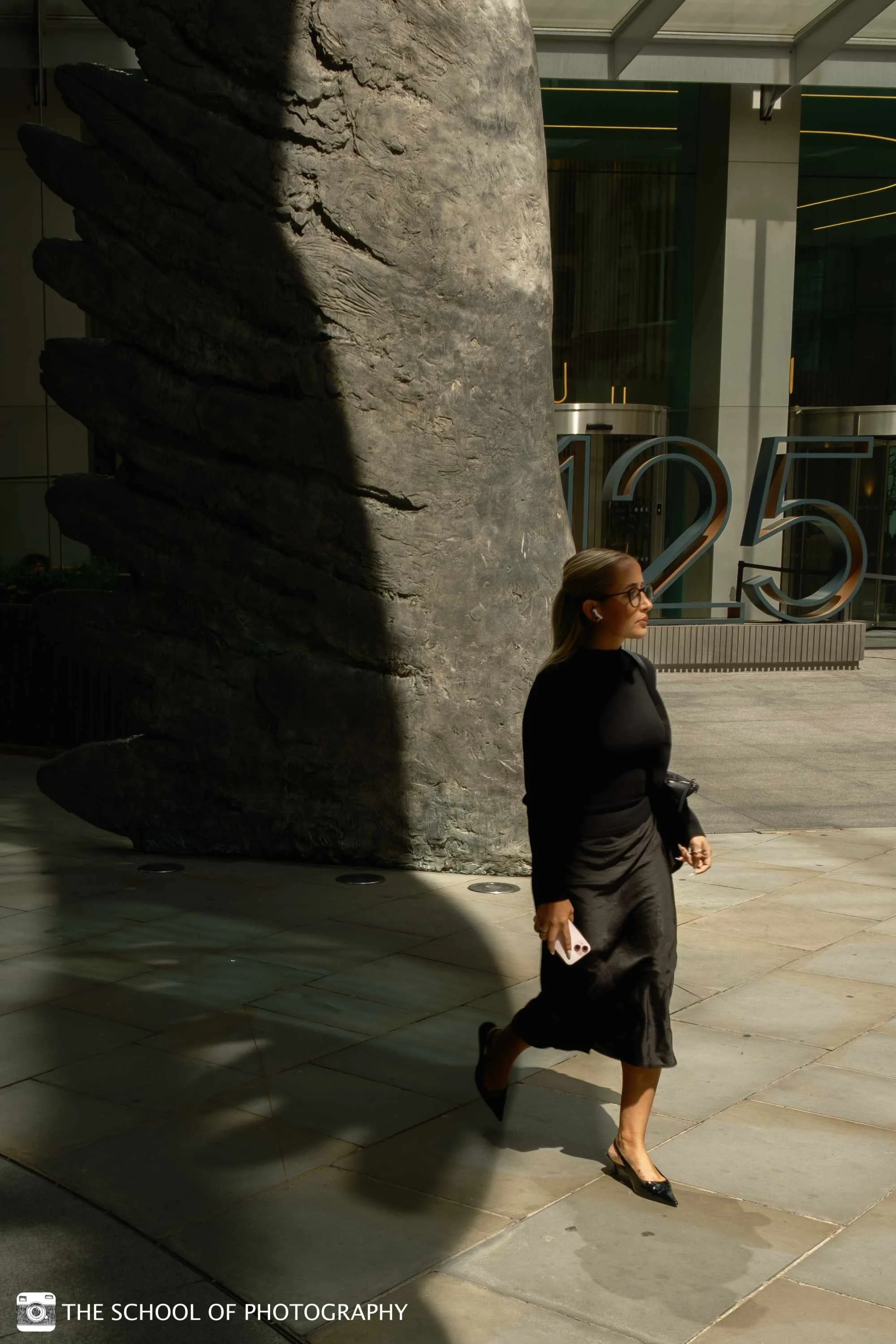 Colour street photography image of a woman walking through strong sunlight beside a large textured sculpture, creating dramatic contrast between light and shadow.