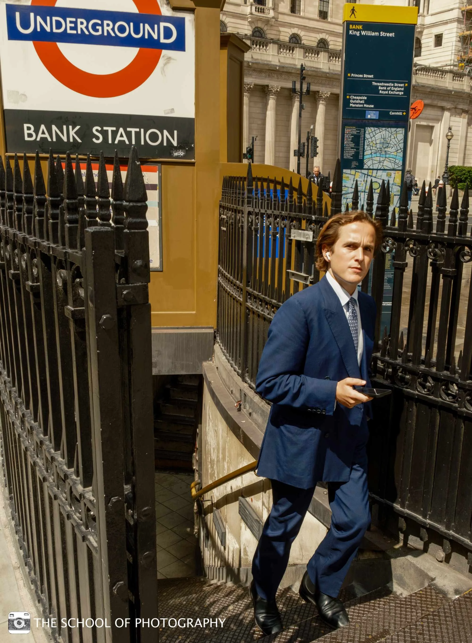 Colour street photography image of a man in a blue suit emerging from Bank Underground station in London, captured in strong sunlight with bold urban lines and contrasting shadows.