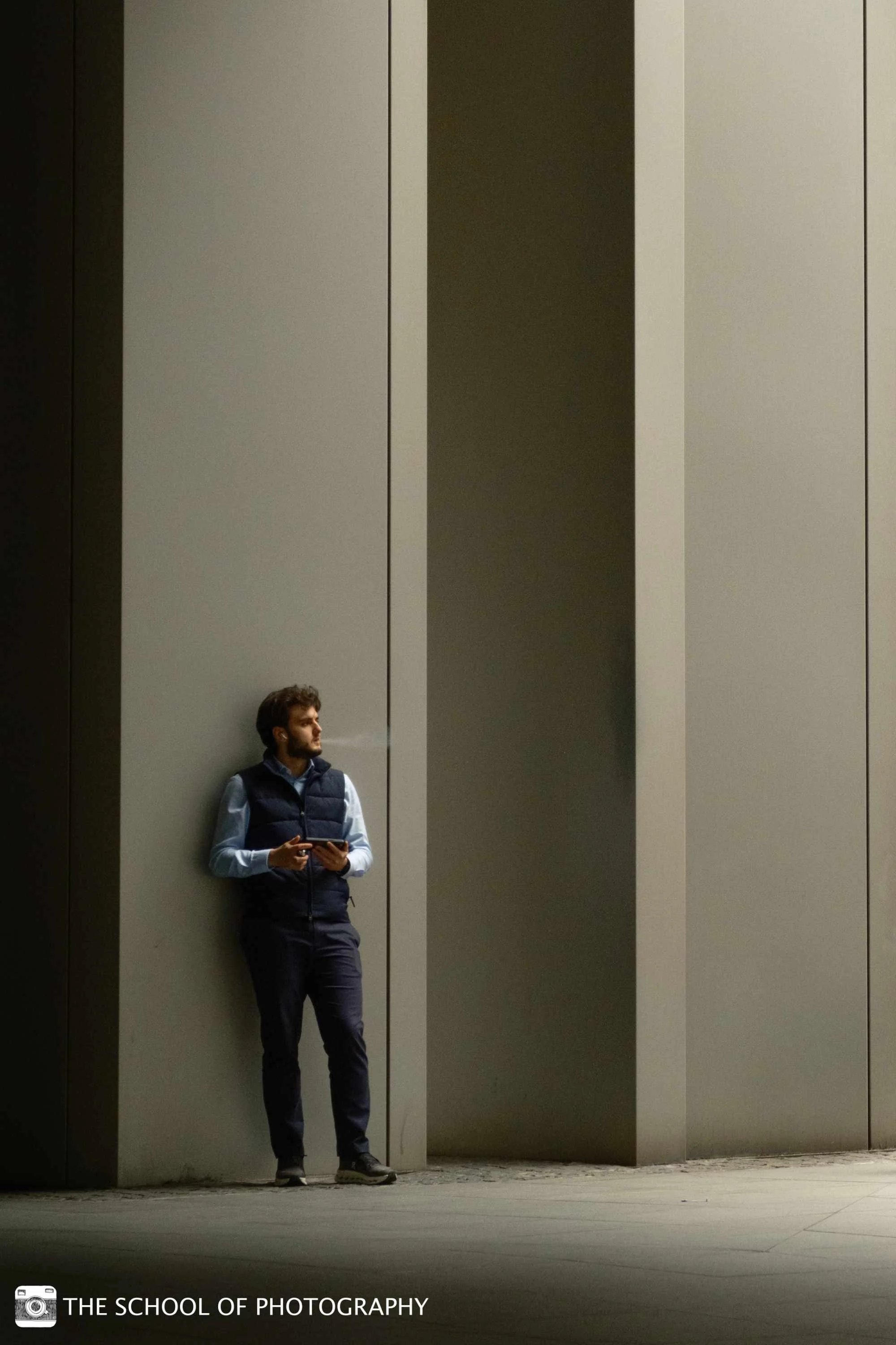 Colour street photography image of a man standing against a modern building, using strong vertical lines, deep shadows and soft light to create a minimalist composition.