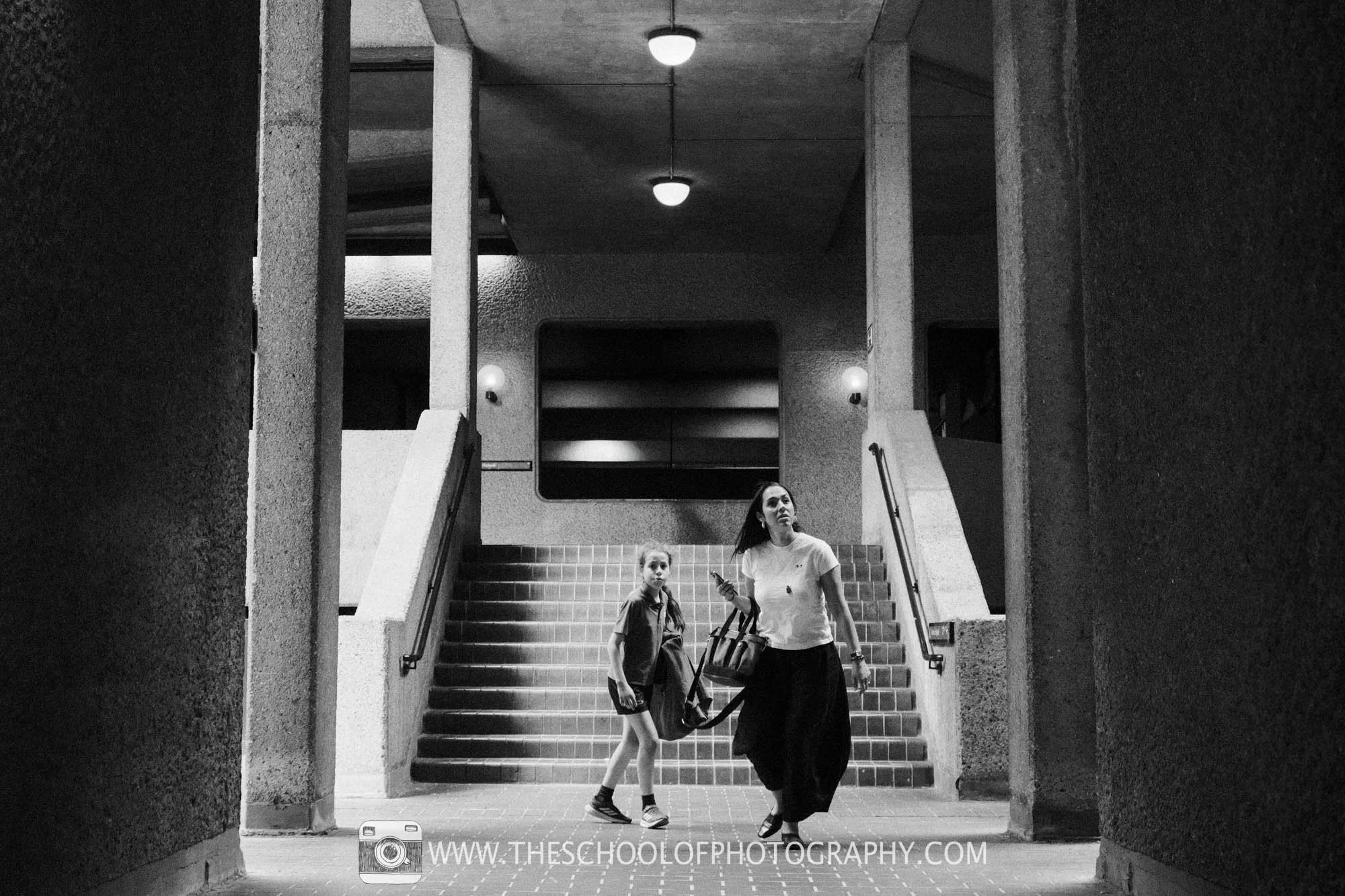 Strong symmetry, repeating lights and Brutalist architecture frame a solitary figure at the end of the walkway.