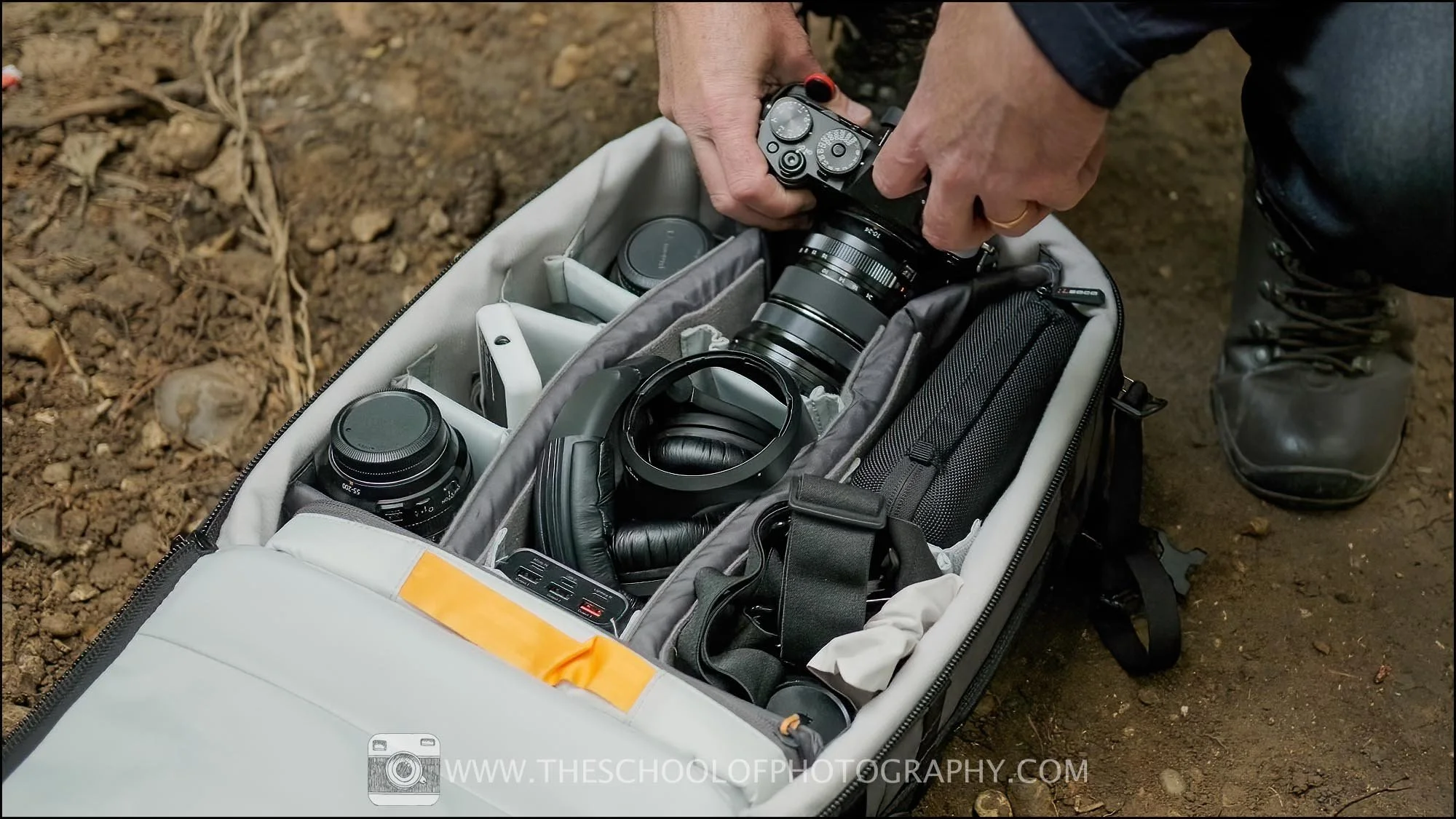 Close-up of a photographer placing a mirrorless camera into a padded camera bag filled with lenses, headphones, and essential videography gear outdoors.