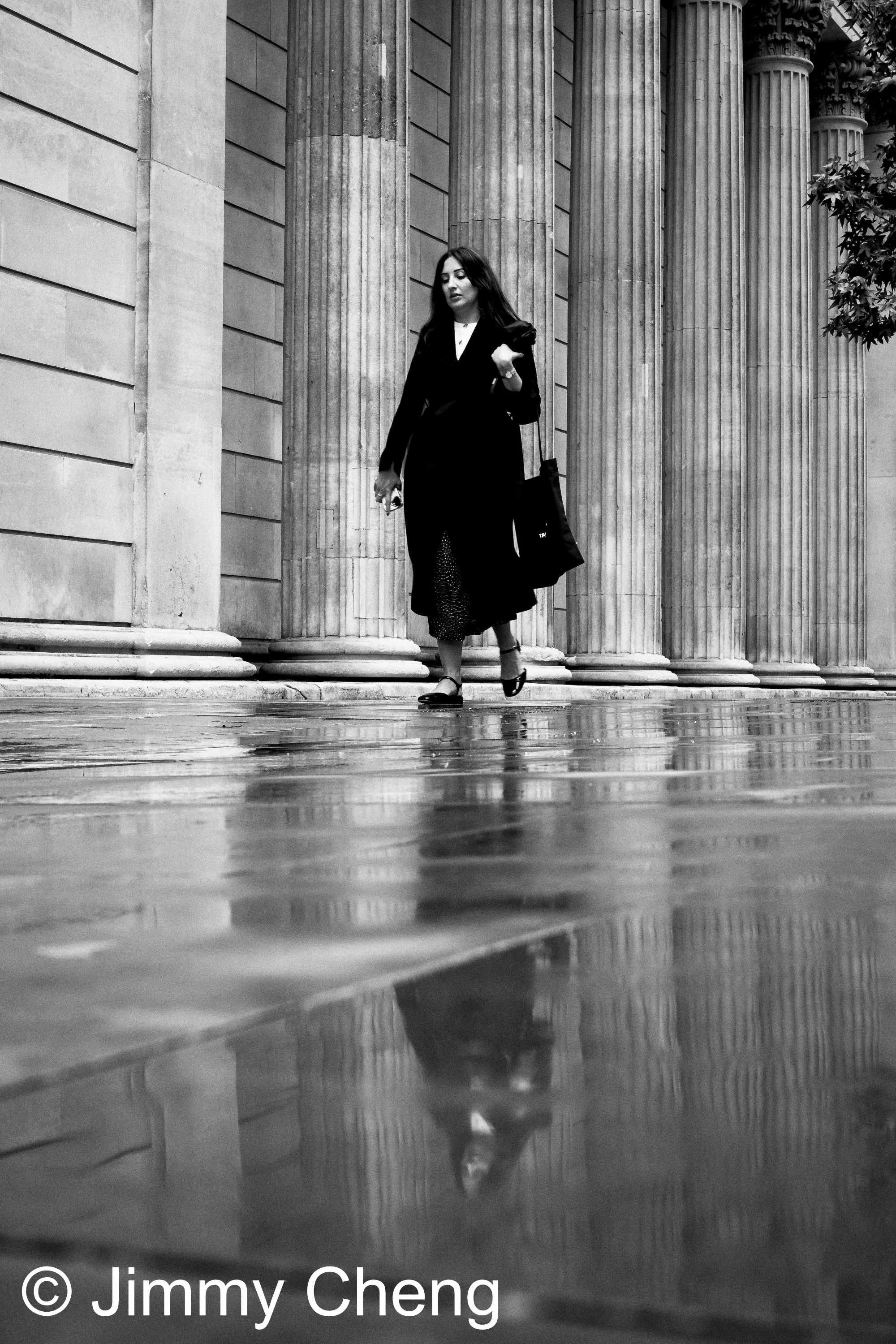 Black and white London street photography showing a woman walking beside classical columns, with her reflection mirrored in the wet pavement after rain.